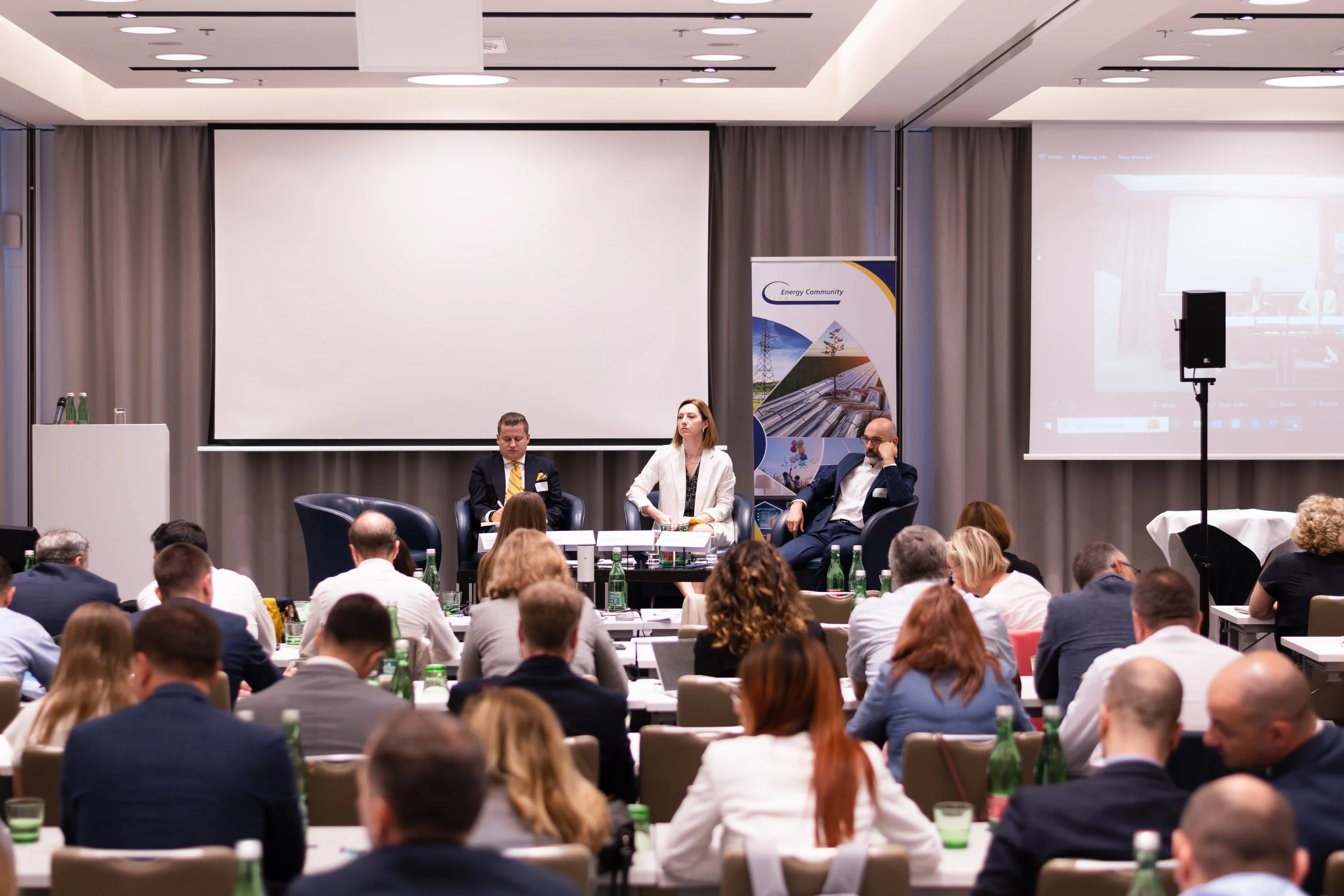 A conference room filled with attendees, three panelists seated at the front, large projection screens on both sides, and water bottles on the tables.