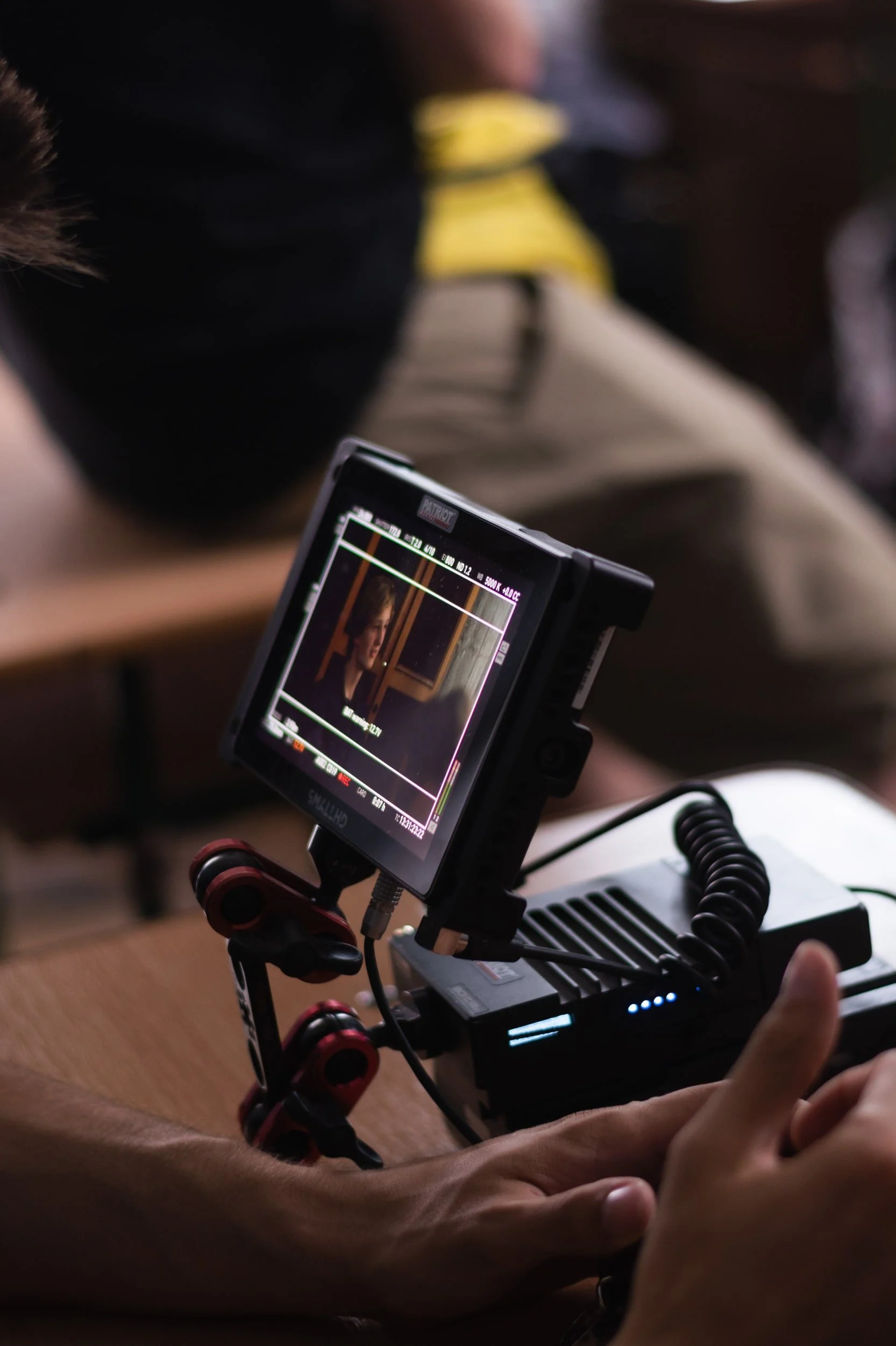 Close-up of a professional camera monitor showing a scene with a woman, mounted on a stabilizer rig held by a person with crossed arms. The background is blurred with a person in a yellow shirt.