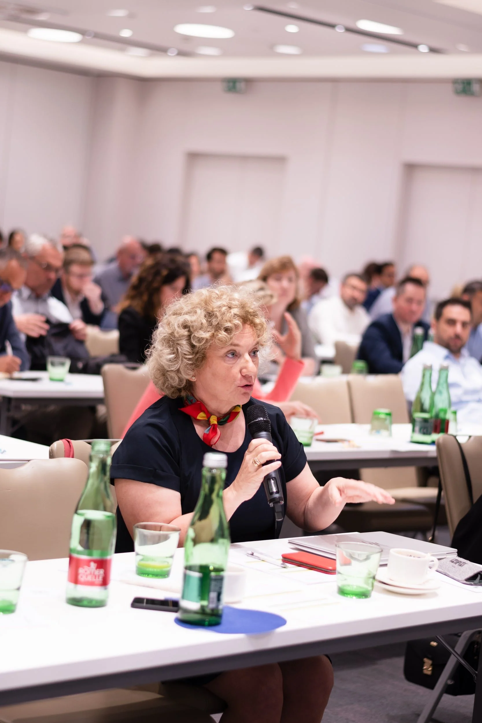 A woman with curly blonde hair and a red and yellow scarf speaking into a microphone during a conference or meeting. She is seated at a table with green bottles of water, glass cups, papers, and a smartphone in front of her. The room is filled with o