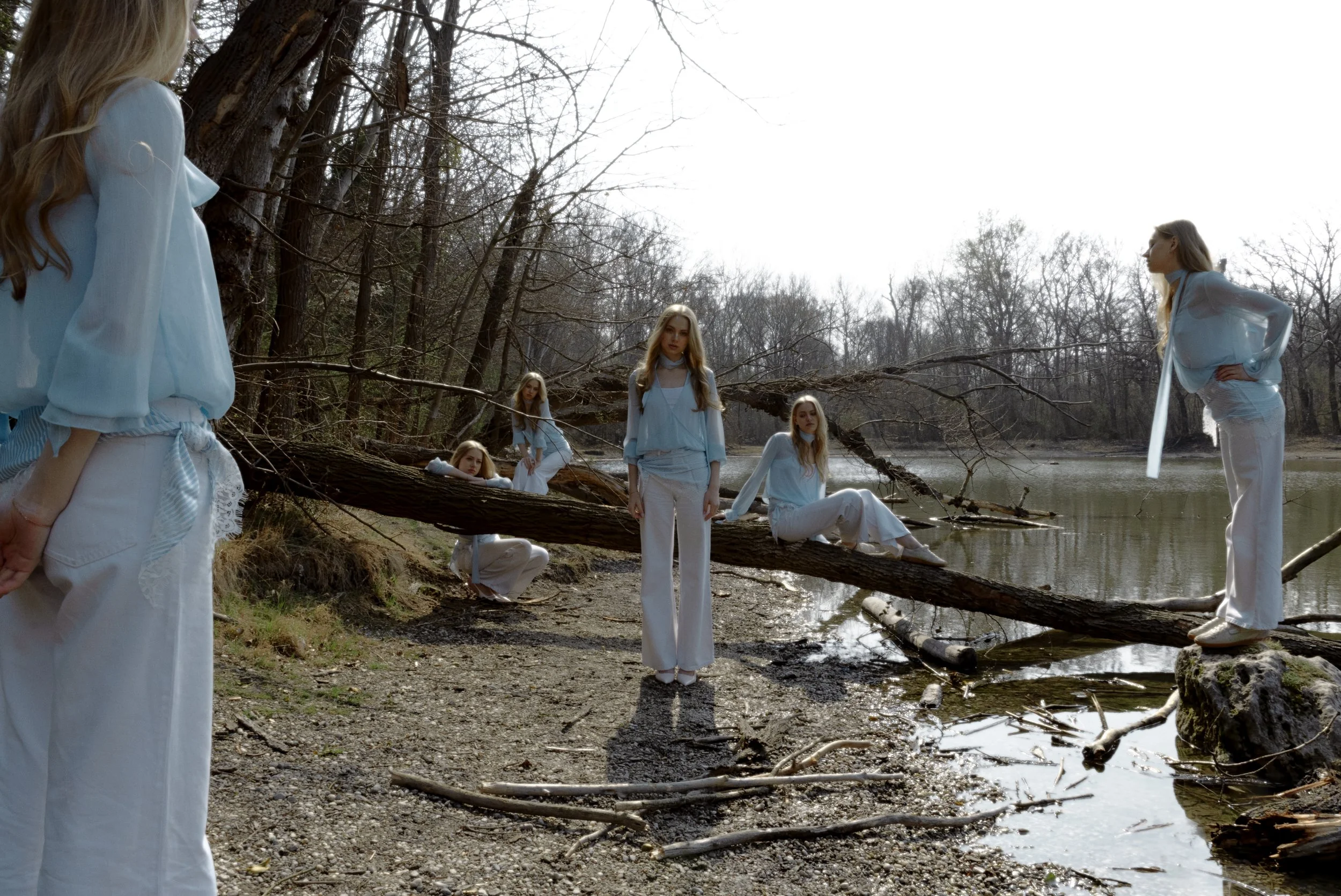Group of women dressed in white and light blue standing, sitting, and leaning on a fallen tree by a river in a wooded area during daytime.