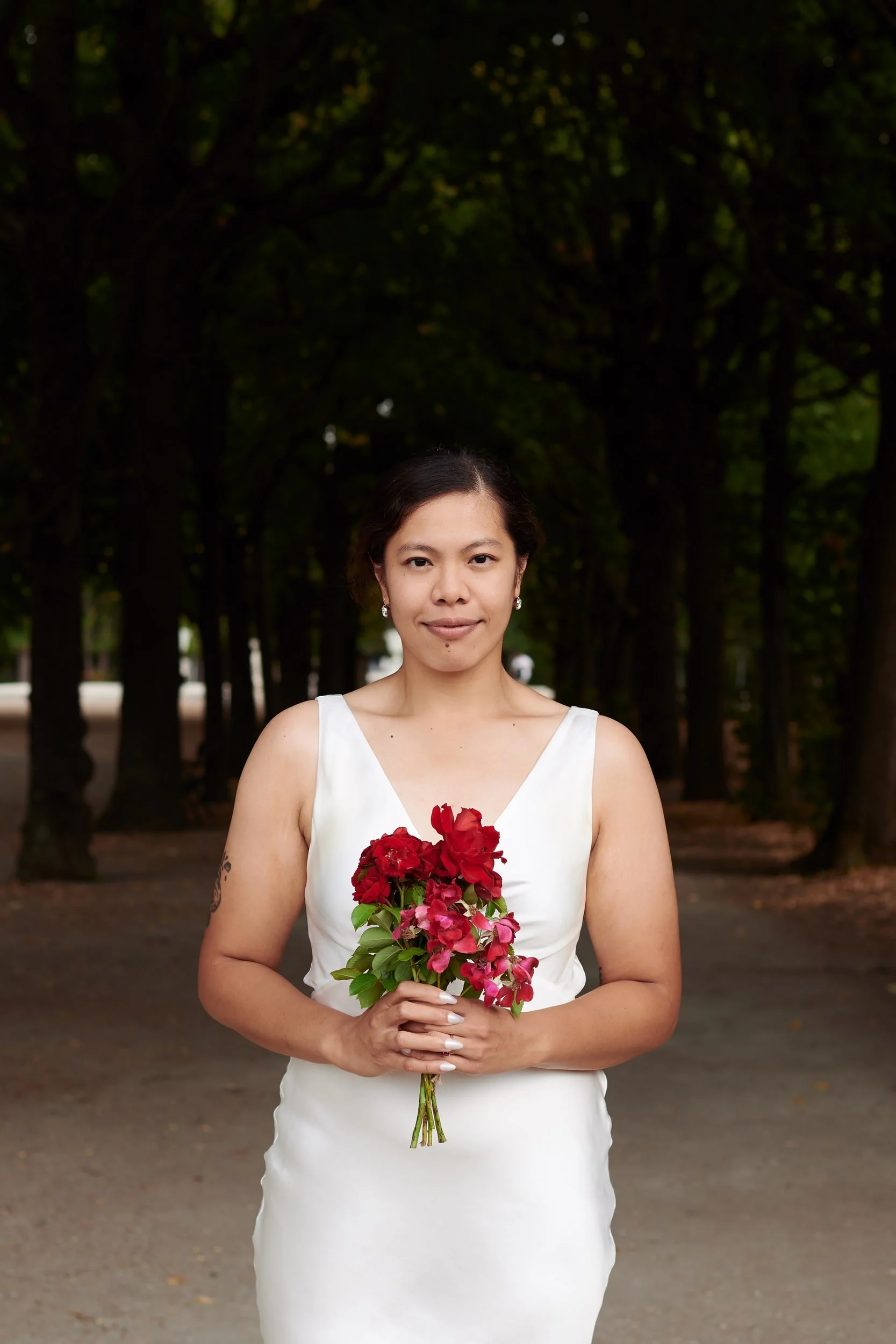 A woman in a white dress holding a bouquet of pink and red flowers, standing outdoors in front of a row of trees.