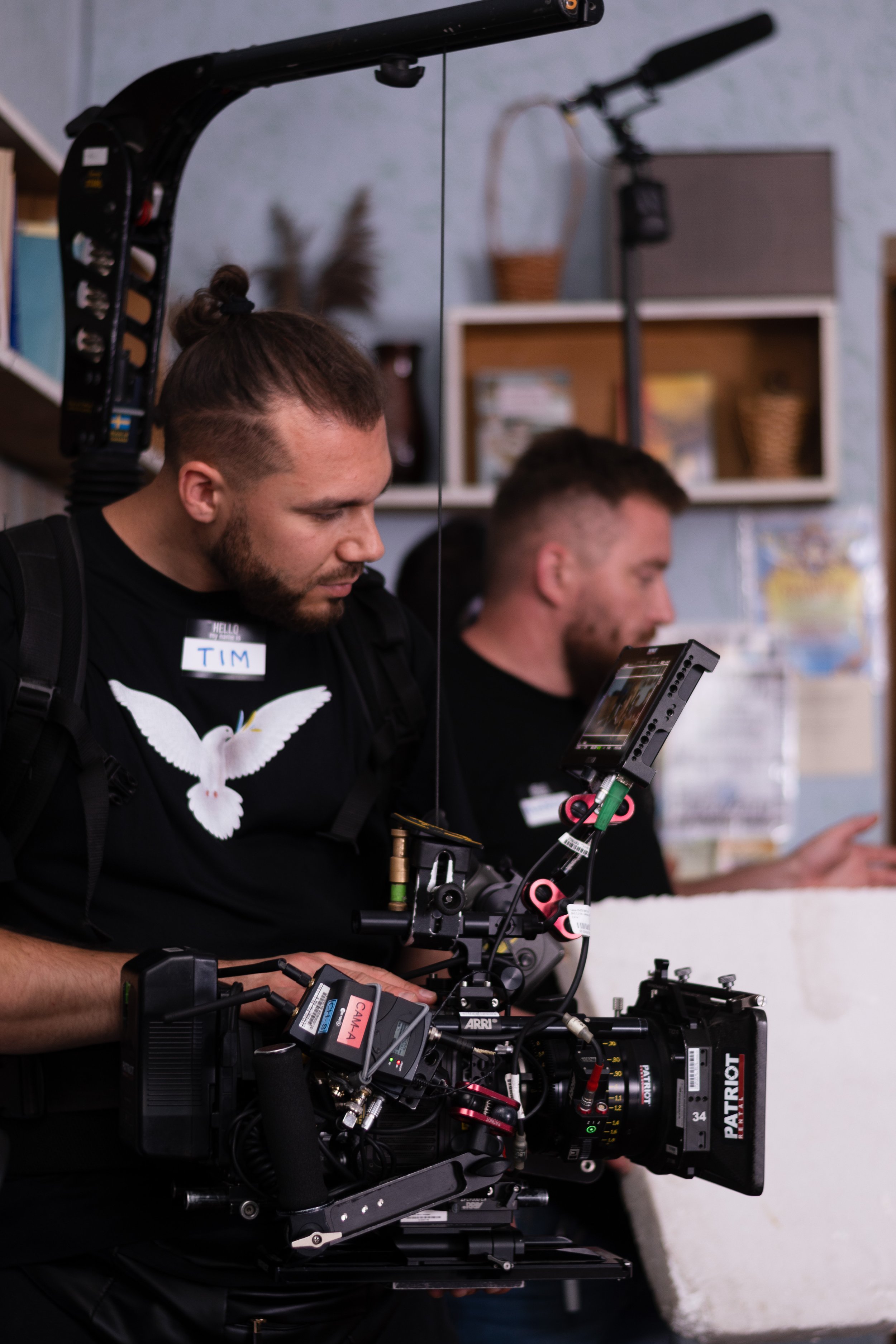 A man operating a professional camera setup with a focus puller and monitor, wearing a black shirt with a white dove graphic and a name tag that says 'Tim'.