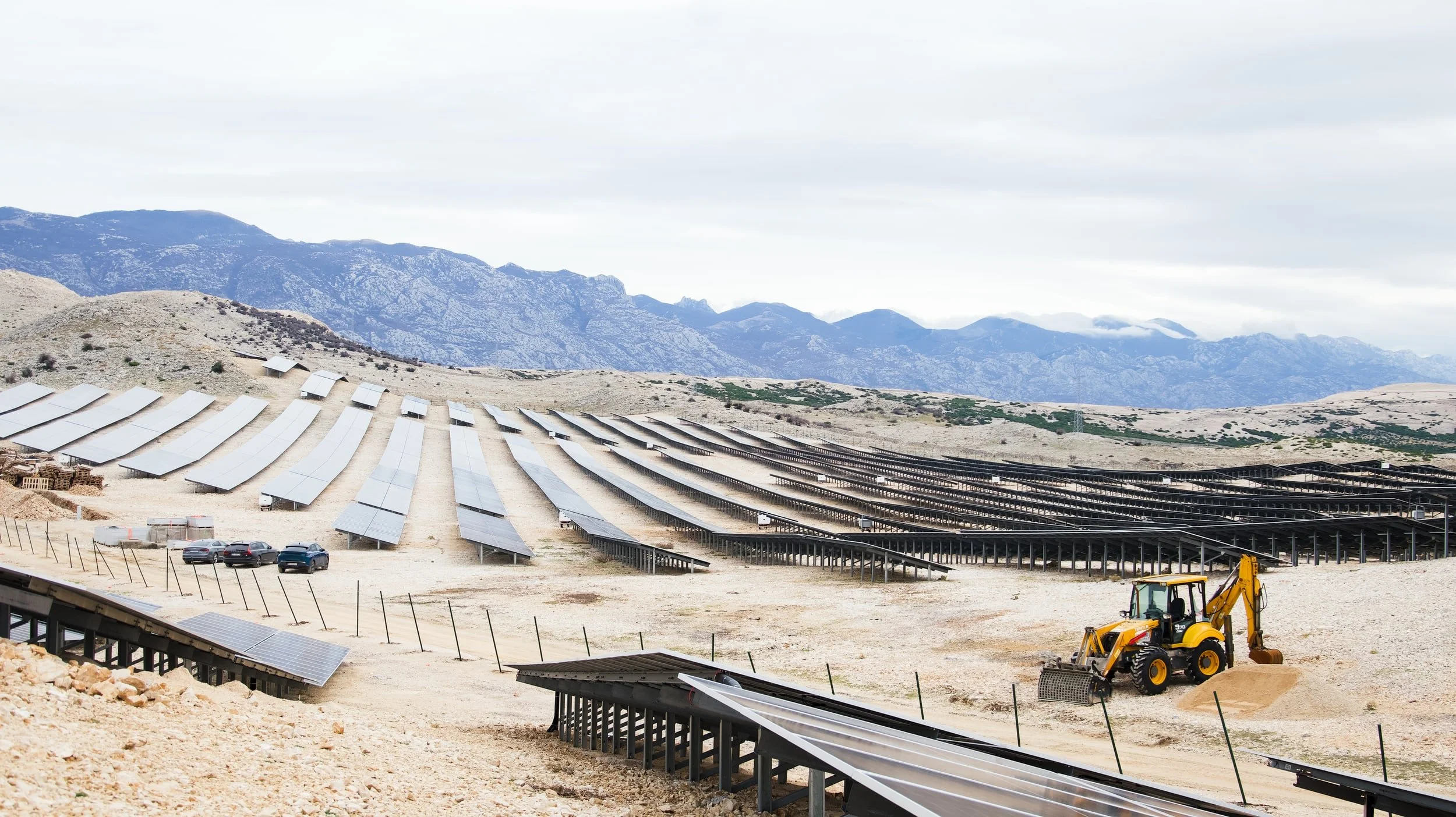 Solar panels installed on the ground in a mountainous area, with a bulldozer nearby, under a cloudy sky.