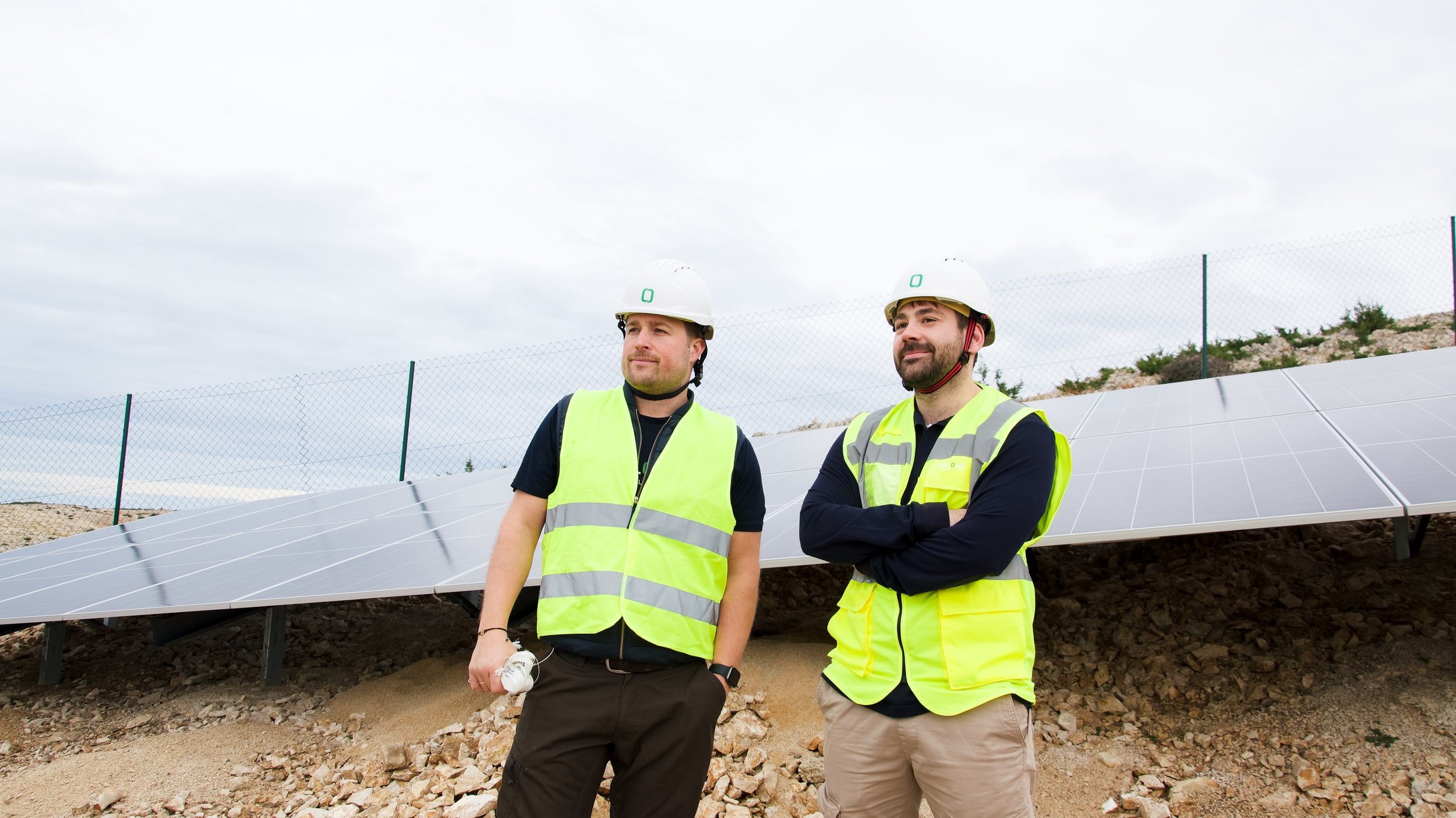 Two men wearing safety helmets and yellow safety vests standing outdoors near solar panels on a cloudy day.