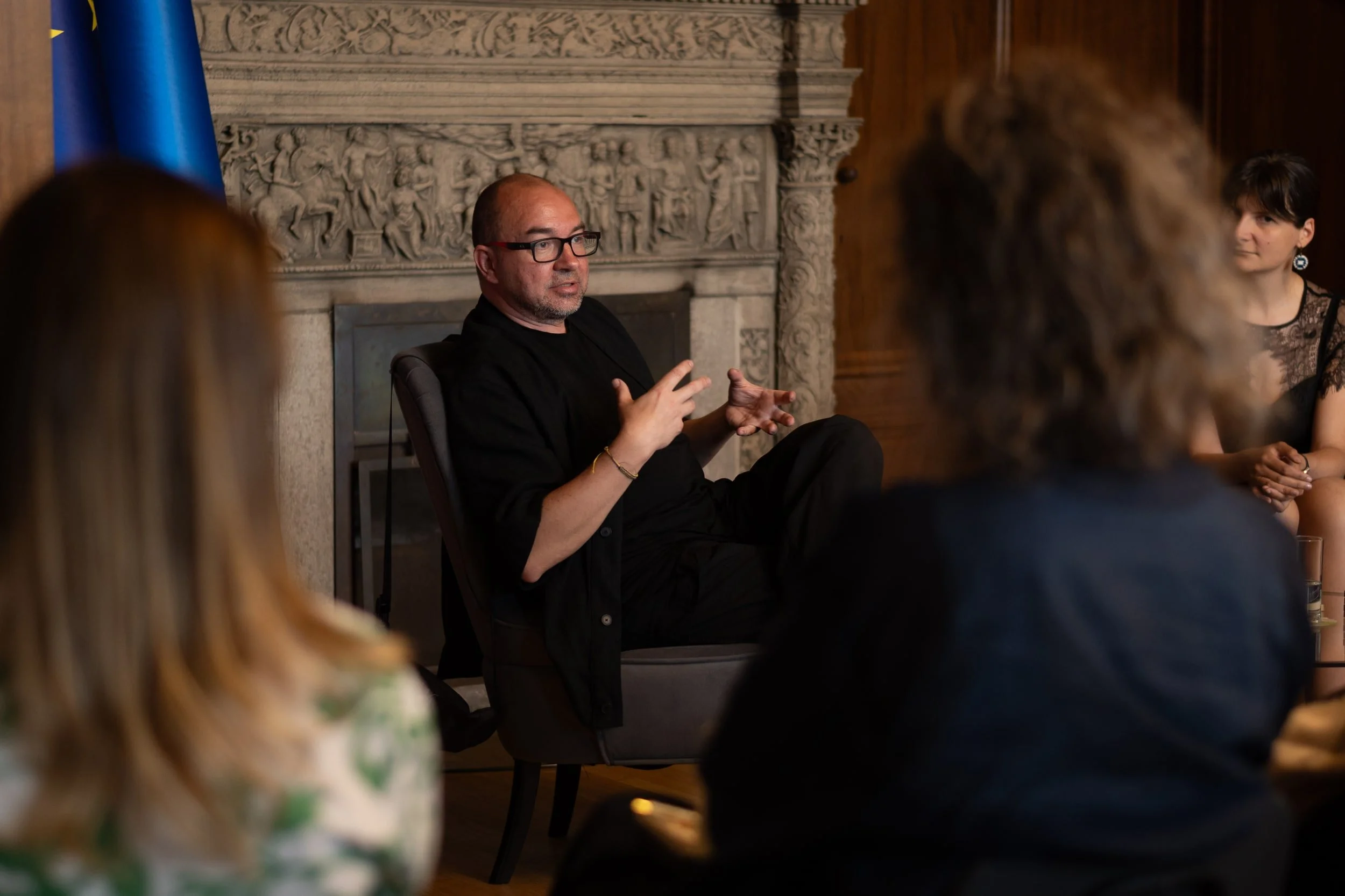 A man with glasses and a beard discusses while seated at a round table during a formal meeting or event, with multiple women listening. They are in a room with a carved stone fireplace and dark wood paneling.