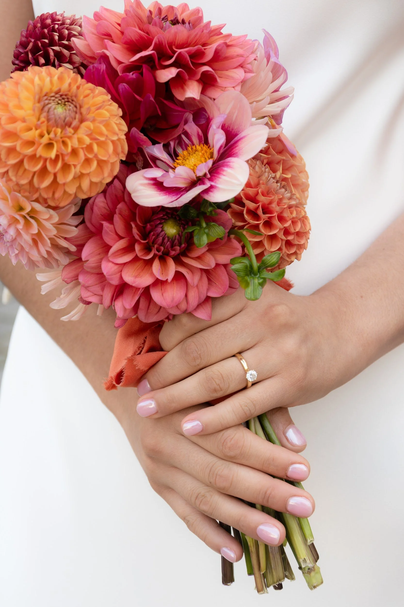 Person holding a bouquet of pink, peach, and purple flowers with their hands, that are adorned with a ring on the left ring finger, against a plain white background.