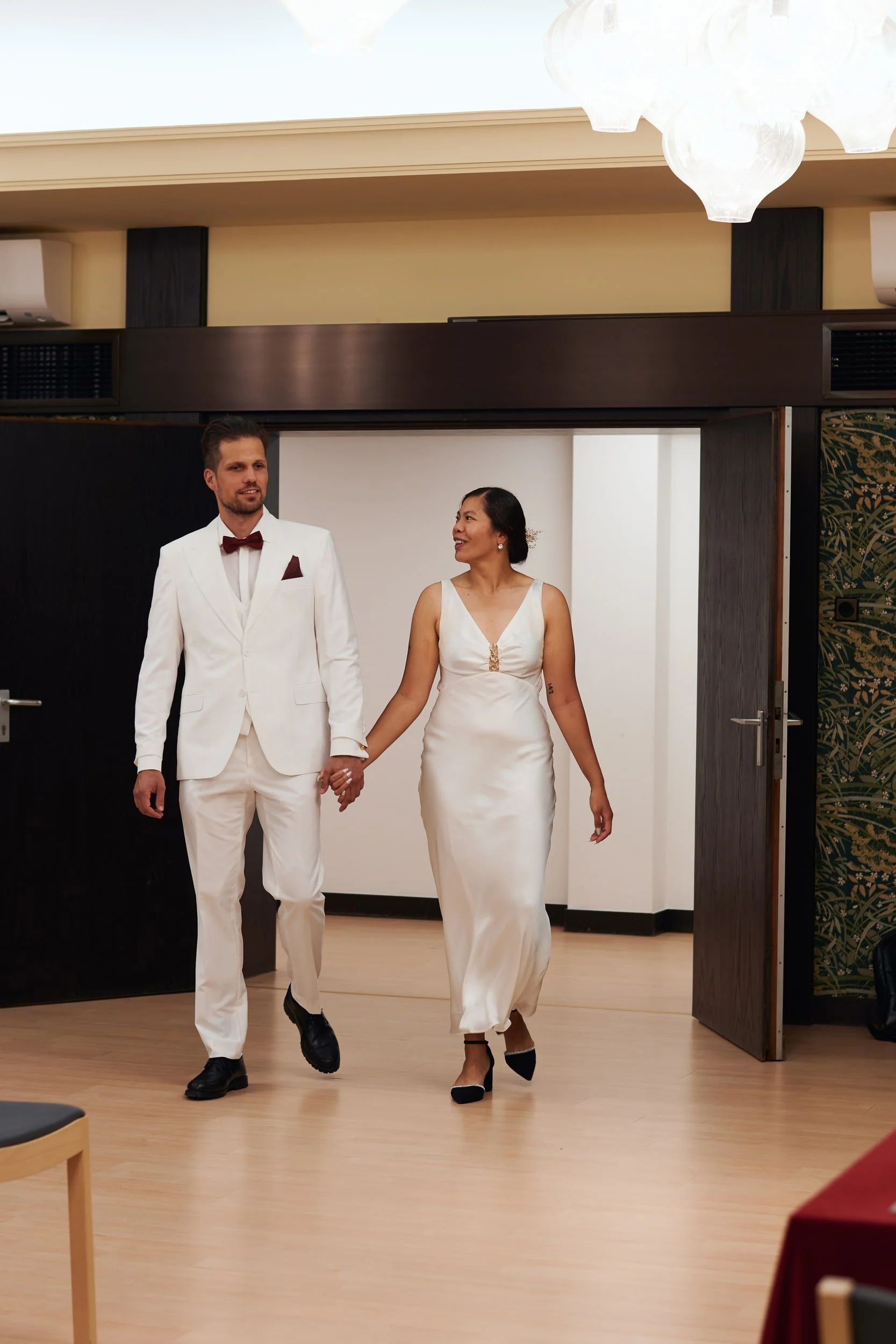 A man and woman in wedding attire walking hand in hand indoors, smiling and looking at each other.