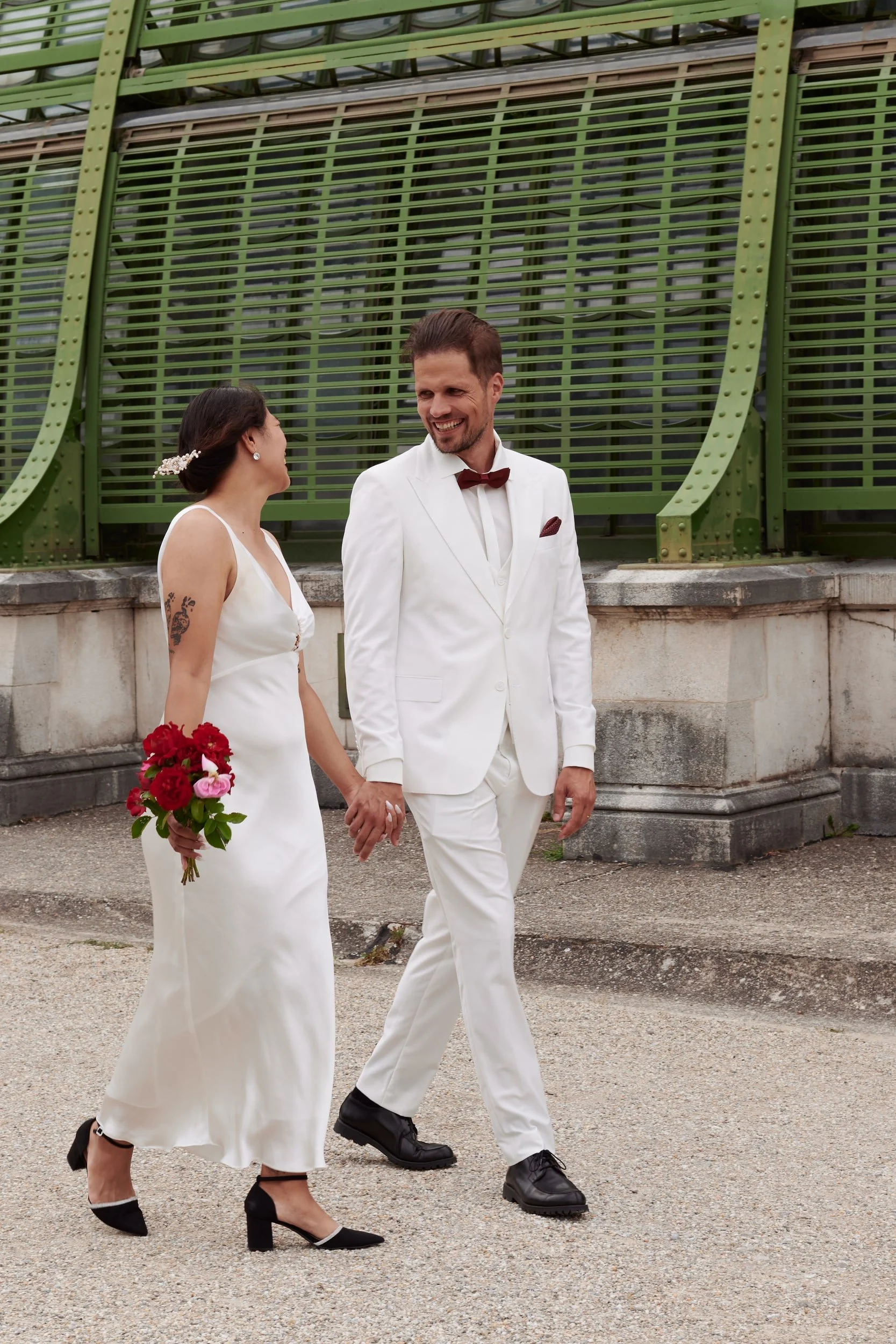 A bride and groom walking hand in hand outdoors near a modern green structure. The bride is wearing a white dress and black high heels, holding a bouquet of red and pink flowers. The groom is dressed in a white suit with a red bow tie and black shoes