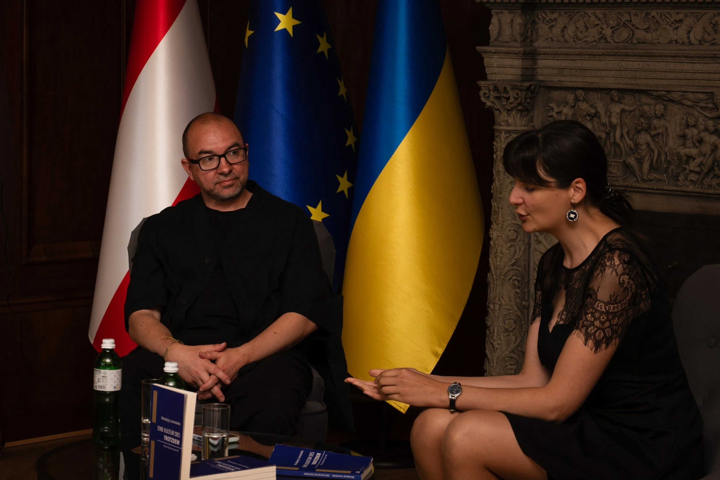 Two individuals, a man and a woman, sitting in front of flags of Austria, the European Union, and Ukraine, engaged in conversation. The setting appears to be a formal meeting or diplomatic event.