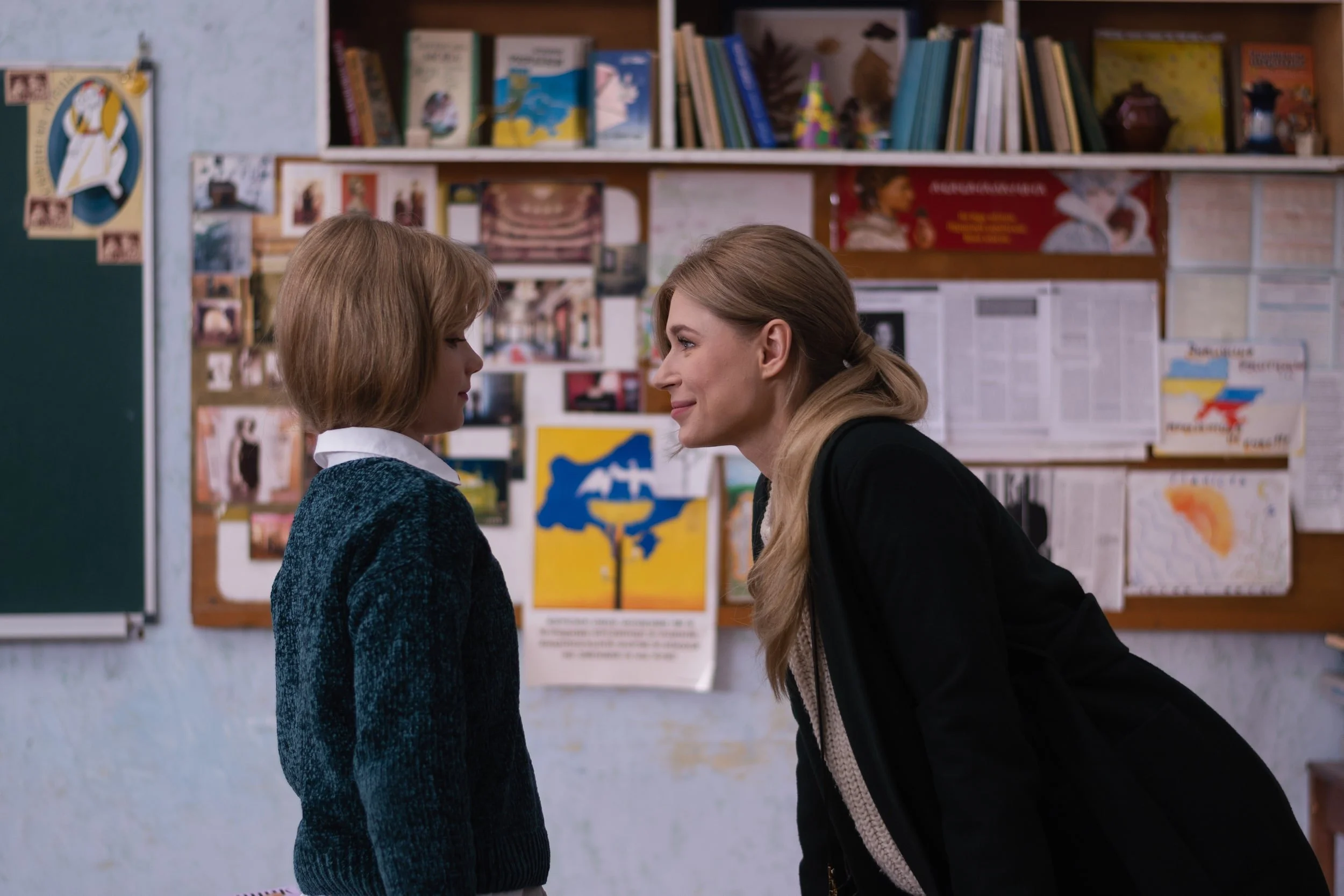 A woman leaning towards a young girl in a classroom, both with short hair, engaging in a close and attentive conversation. The background features a bulletin board with various posters, books, and artwork.