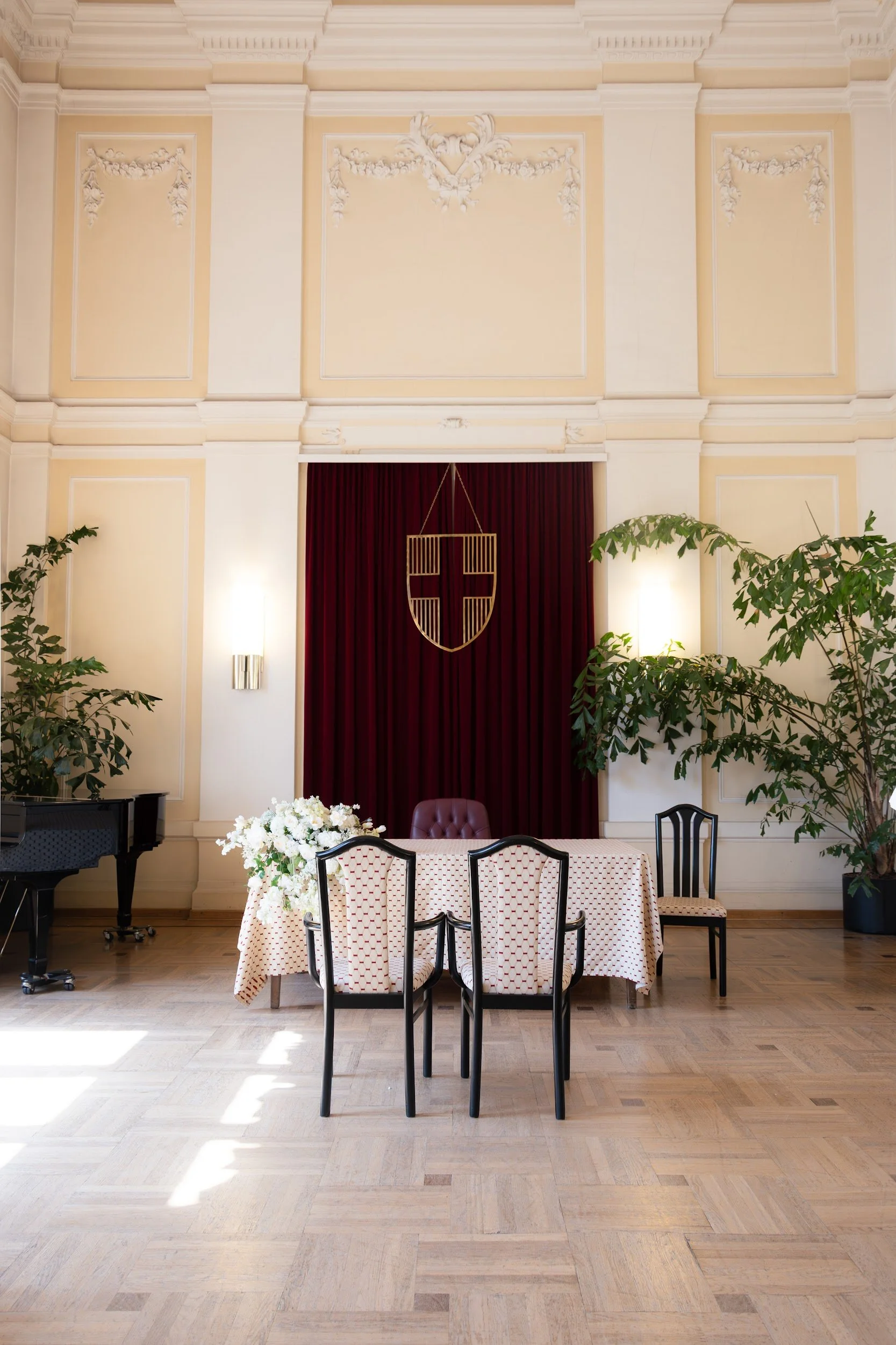 A formal dining or conference room with a round table covered with a red, white, and black checkered tablecloth, surrounded by black and beige chairs. A floral centerpiece is on the table. Behind the table, there is a red velvet curtain with a gold a