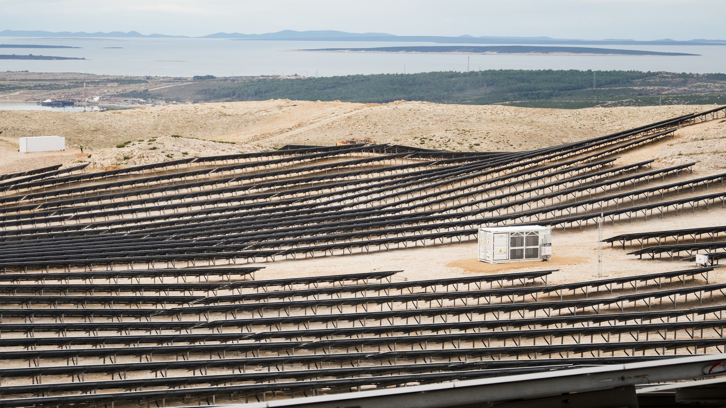 Solar panel installation site on a barren, hilly landscape with distant water and mountain views in the background.