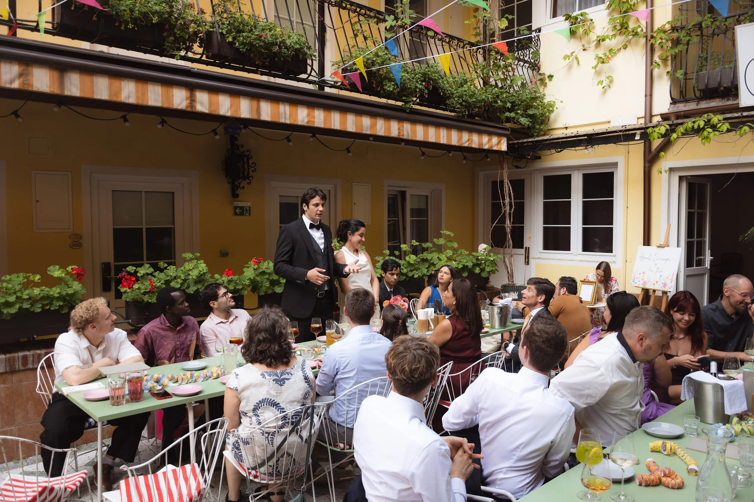 A large group of people gathered around a long table at an outdoor celebration or party, with some standing and others seated, under a balcony decorated with colorful bunting flags and greenery. A man in a tuxedo appears to be speaking or making a to