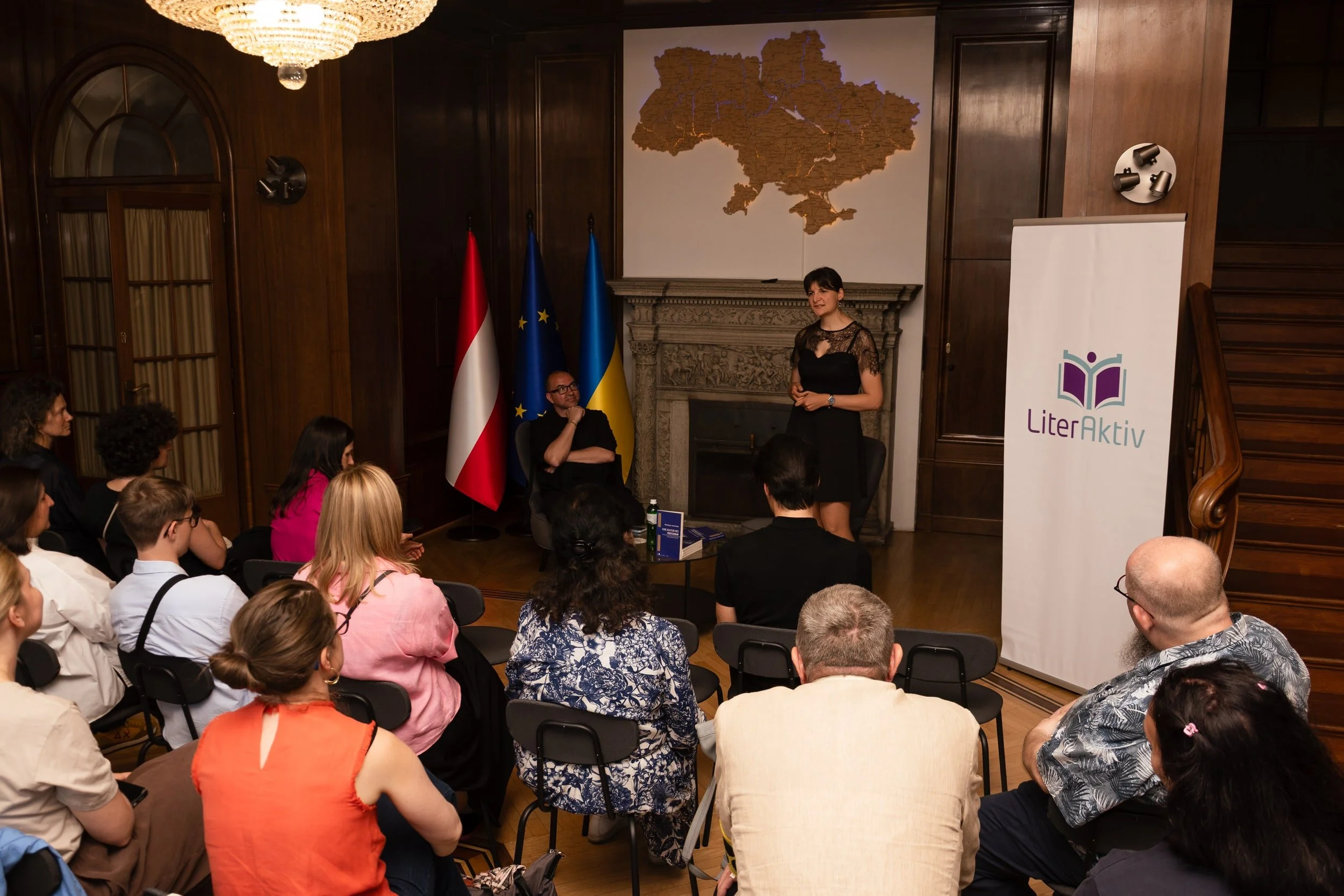 A woman in a black dress giving a presentation to an audience seated in a wood-paneled room with flags of Austria, the European Union, and Ukraine behind her. A map of Ukraine is displayed on a screen above the fireplace.