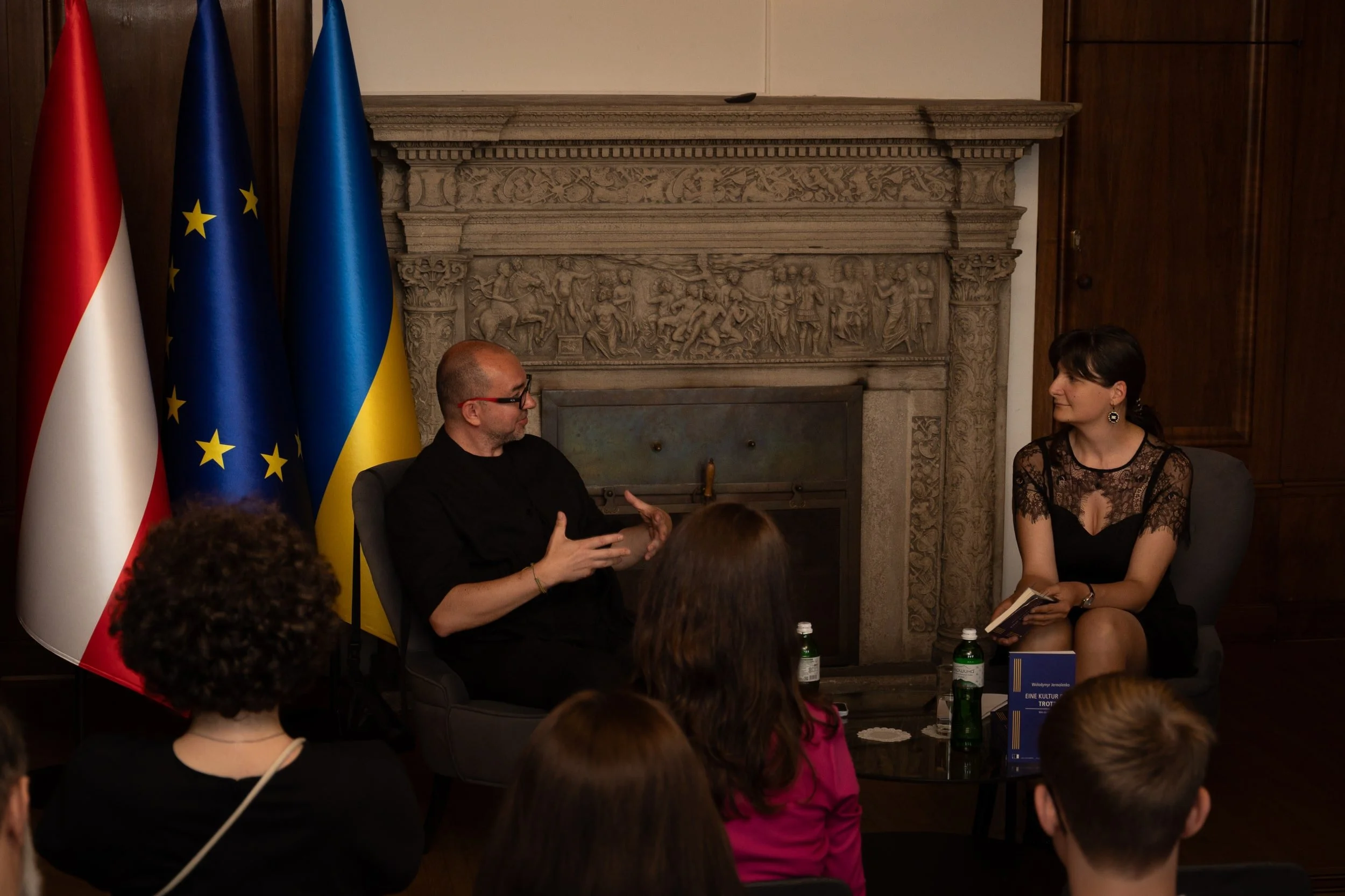 Two people sitting and talking in front of flags, one with a bald head and glasses and the other with dark hair, in a room with ornate stone fireplace and wooden paneling.