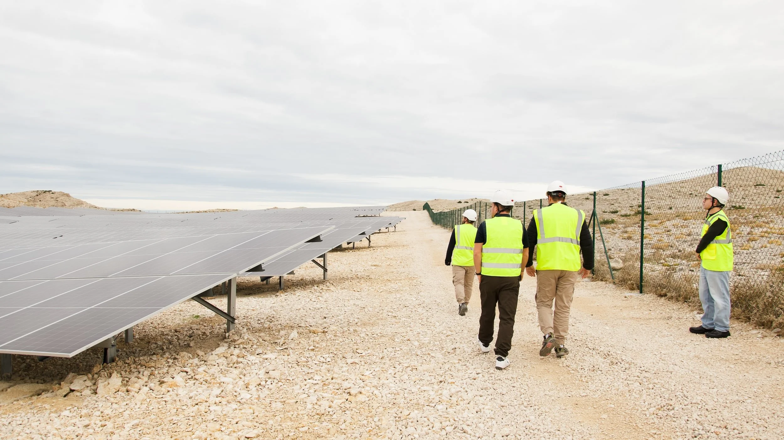 Group of five people in safety vests and helmets walking near solar panels in a desert.