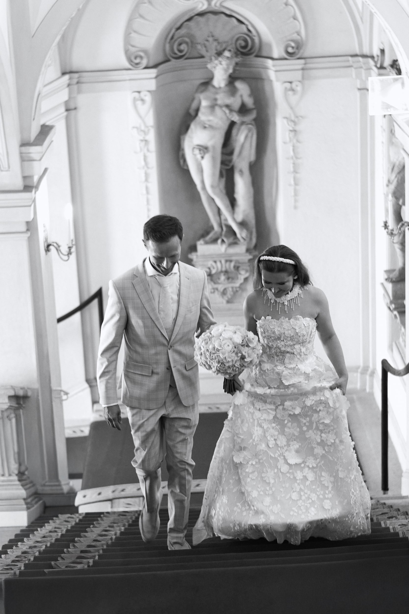 A black and white photo of a bride and groom walking down a church aisle. The bride is wearing a lace wedding gown and holding a large bouquet of flowers. The groom is in a light-colored suit. They are smiling and looking down as they descend the stairs. Behind them, there is a large sculpture of a religious figure in a niche of the ornate church interior.