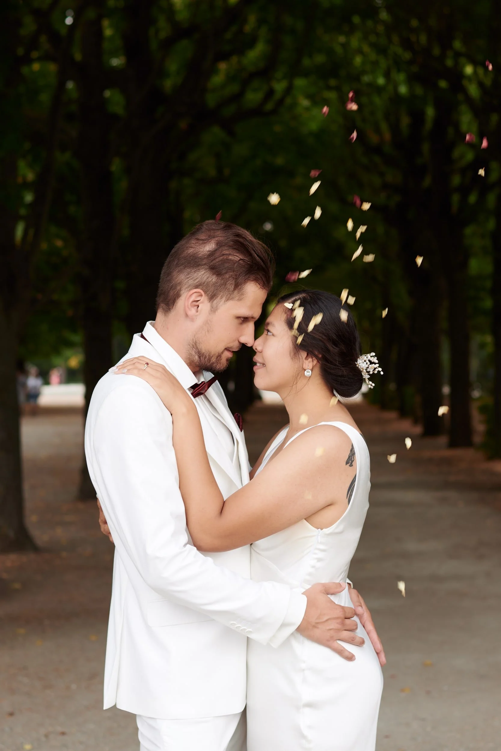 A newlywed couple in white attire embracing each other in a park with dark green trees in the background, surrounded by falling flower petals.