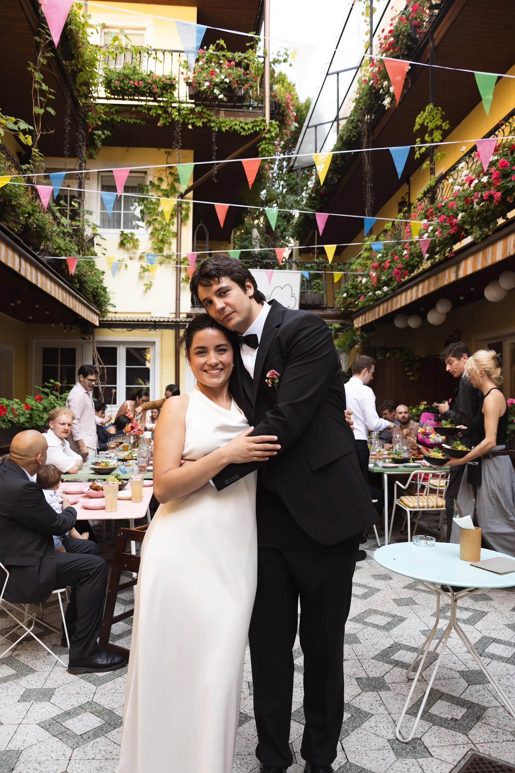 A bride and groom hugging at an outdoor celebration with colorful bunting, tables with food, and other guests in the background.