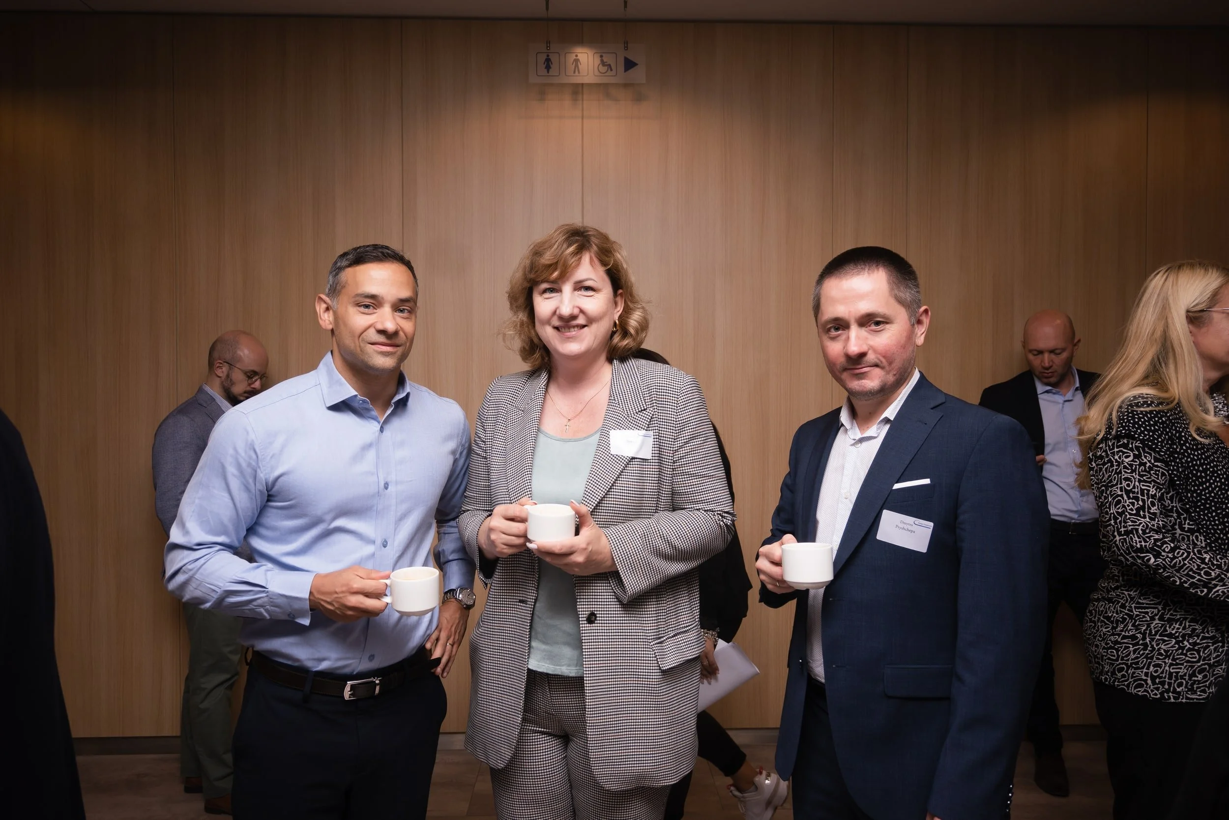 People at a professional networking event holding coffee cups, standing in front of a wooden wall with restroom signs above.