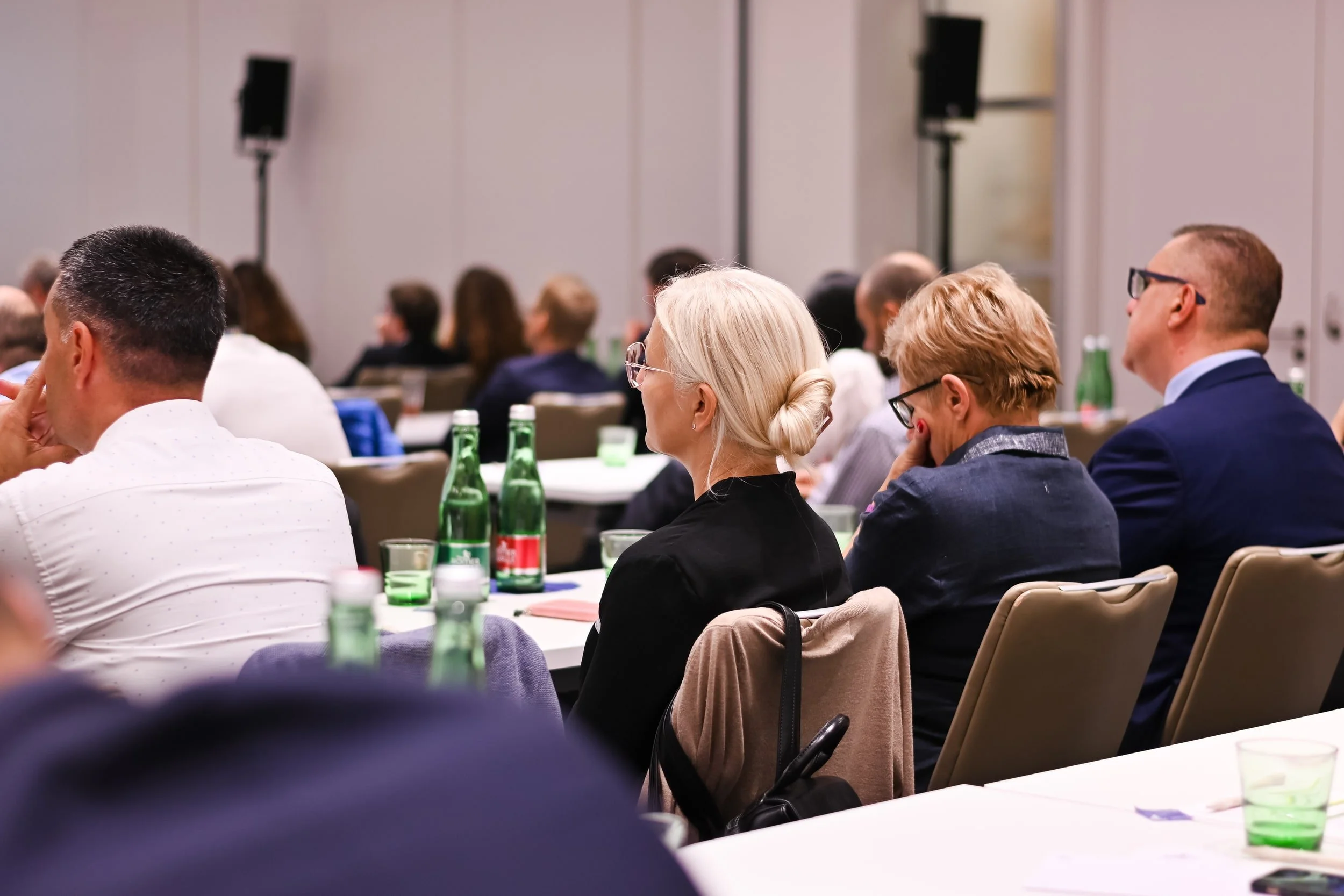 Attendees at a conference or seminar sitting at tables with green bottles of beverages in front of them, listening to a presentation in a large, modern room.