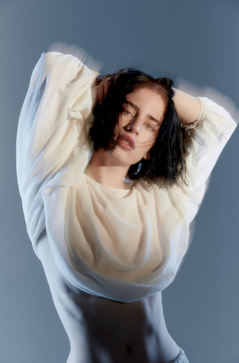 A woman with dark, wavy hair and a nose ring poses with her arms raised above her head, wearing a loose, sheer white top against a plain gray background.