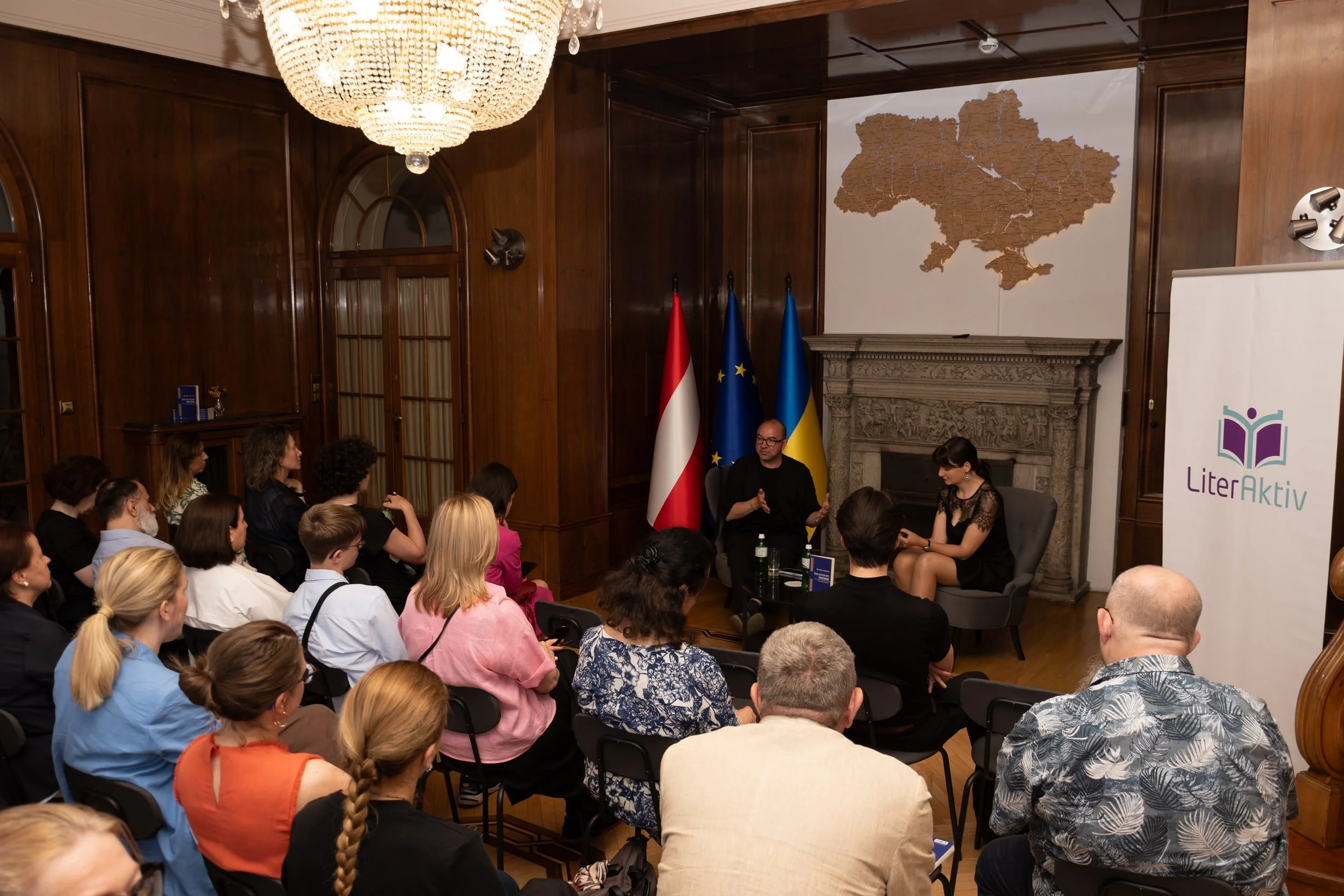 A group of people attending a presentation or panel discussion in a wood-paneled room with flags of Austria, the European Union, and Ukraine behind the speakers. There is a large map of Ukraine on the wall and a banner with the logo 'LiterAktiv'.
