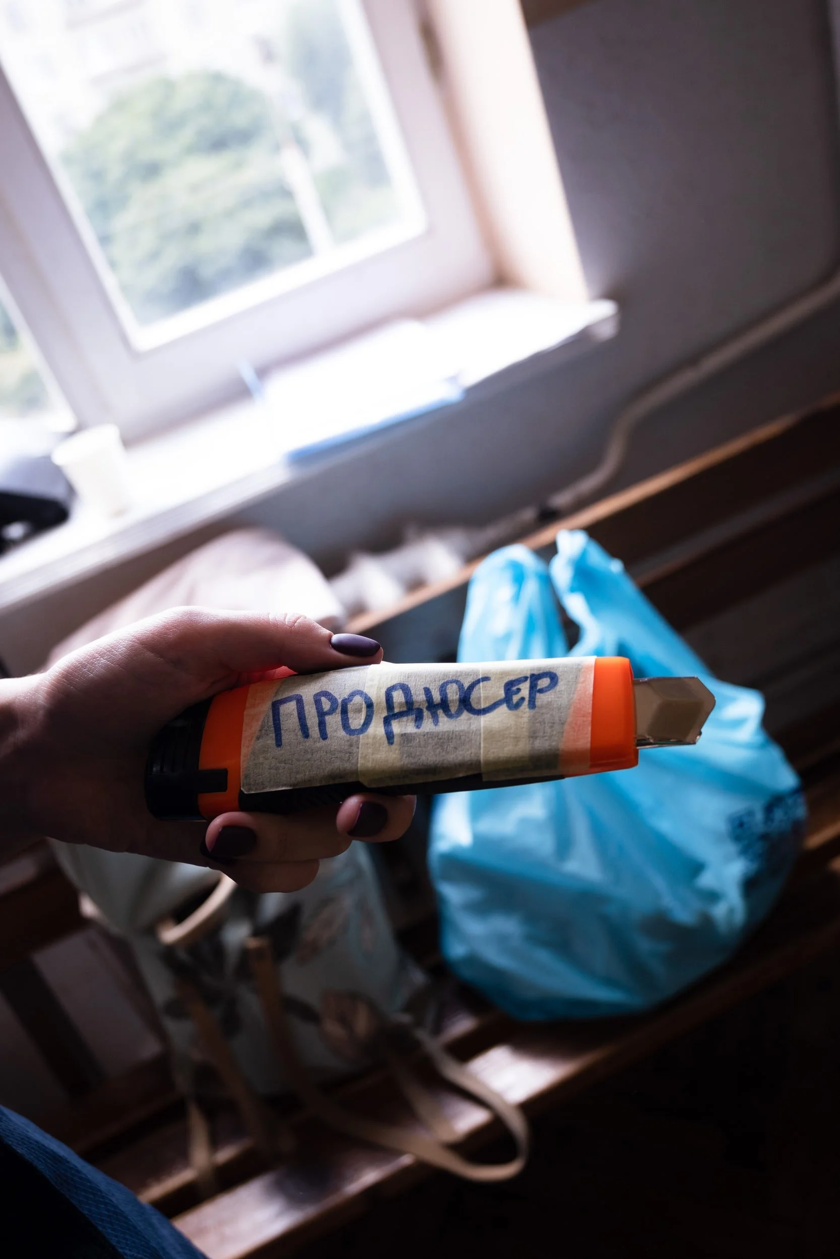 A person's hand holding a small container with handwritten Cyrillic text, with a wooden window sill and a blue plastic bag in the background.