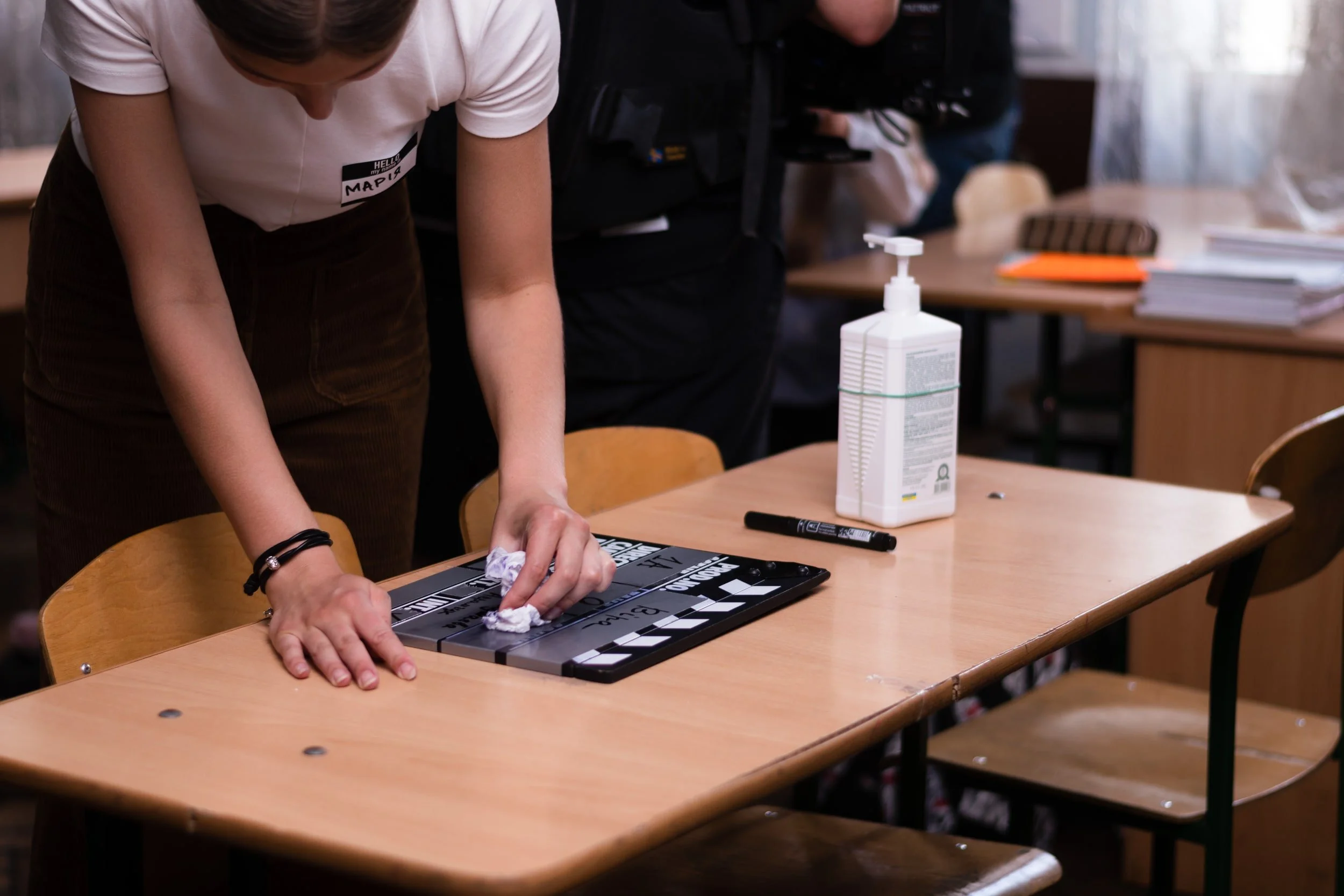 A person cleaning a black and white clapperboard on a wooden table, with a bottle of hand sanitizer nearby in a room with other people and tables.