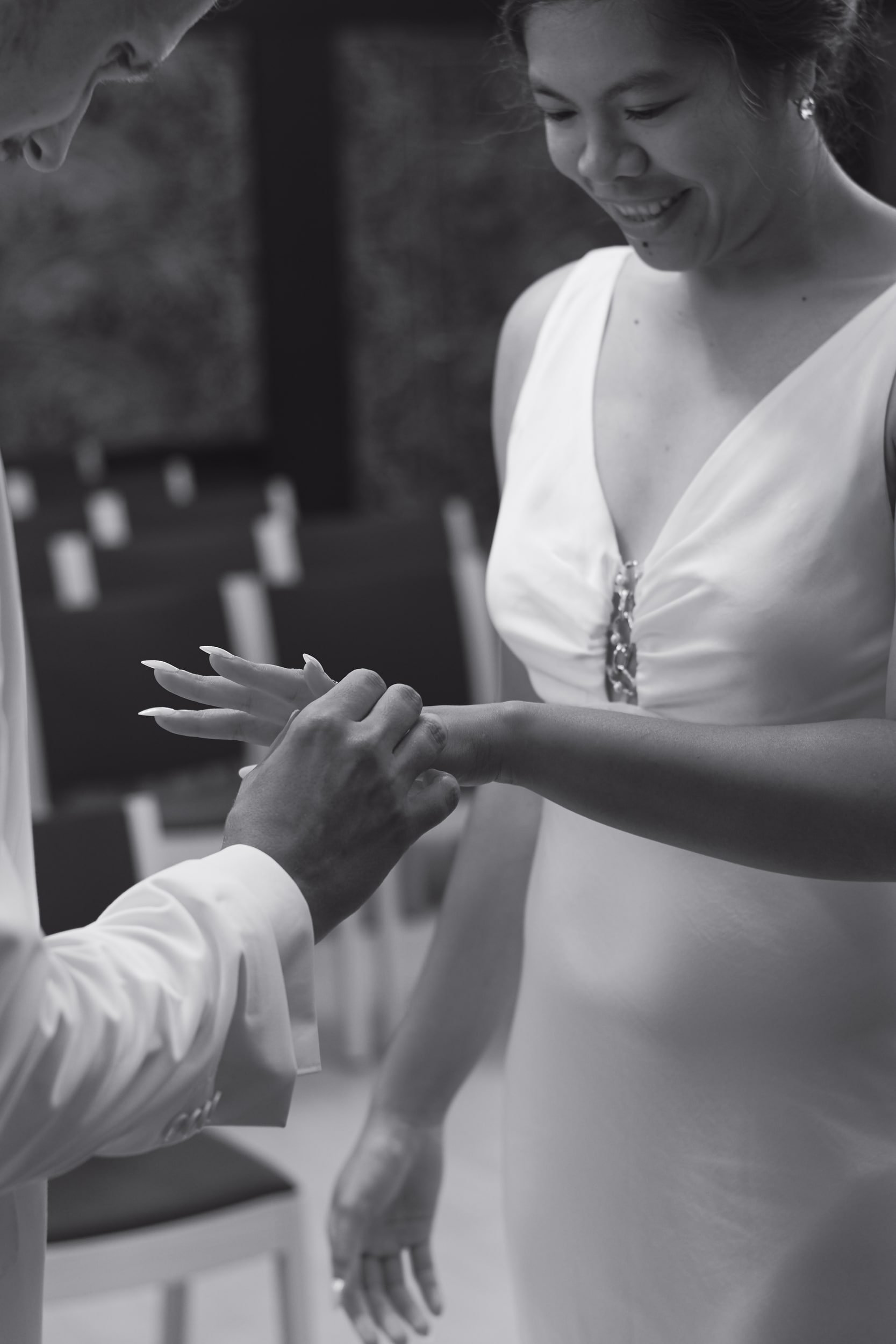 A woman with dark hair tied back, wearing a sleeveless white dress and earrings, is smiling and looking down at her left hand. A man in a white shirt is gently holding her left hand, possibly fitting a ring. The background suggests an outdoor setting