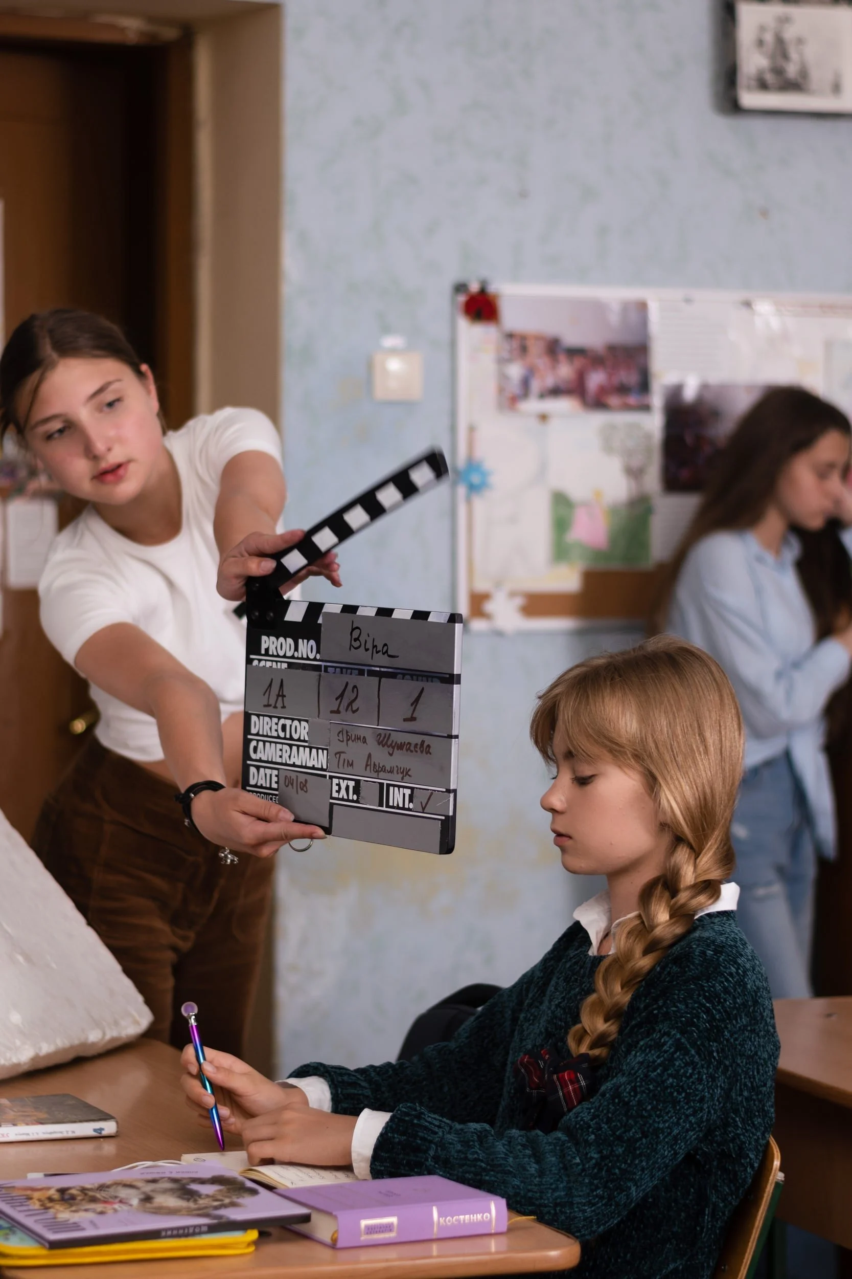 A woman holding a clapperboard labeled 'Bina' in a classroom with students. One student with braided hair is sitting at a desk, writing with a pink pen, while other students are in the background.