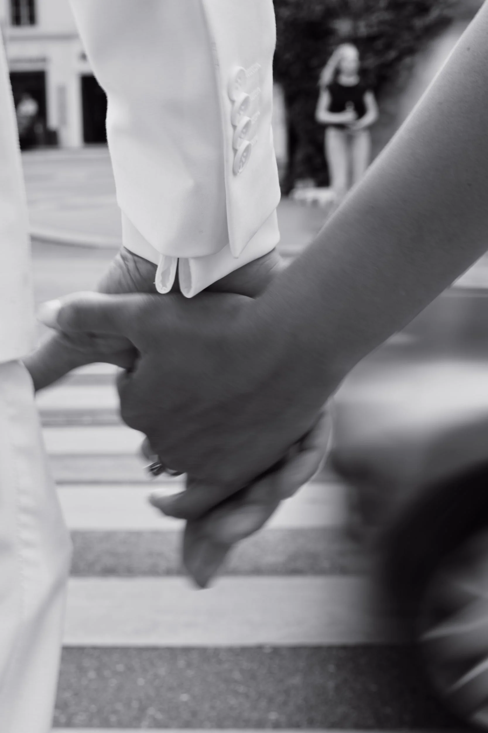 Close-up of a person's hand holding another person's hand, with a person in the background wearing a black shirt and shorts standing outdoors.