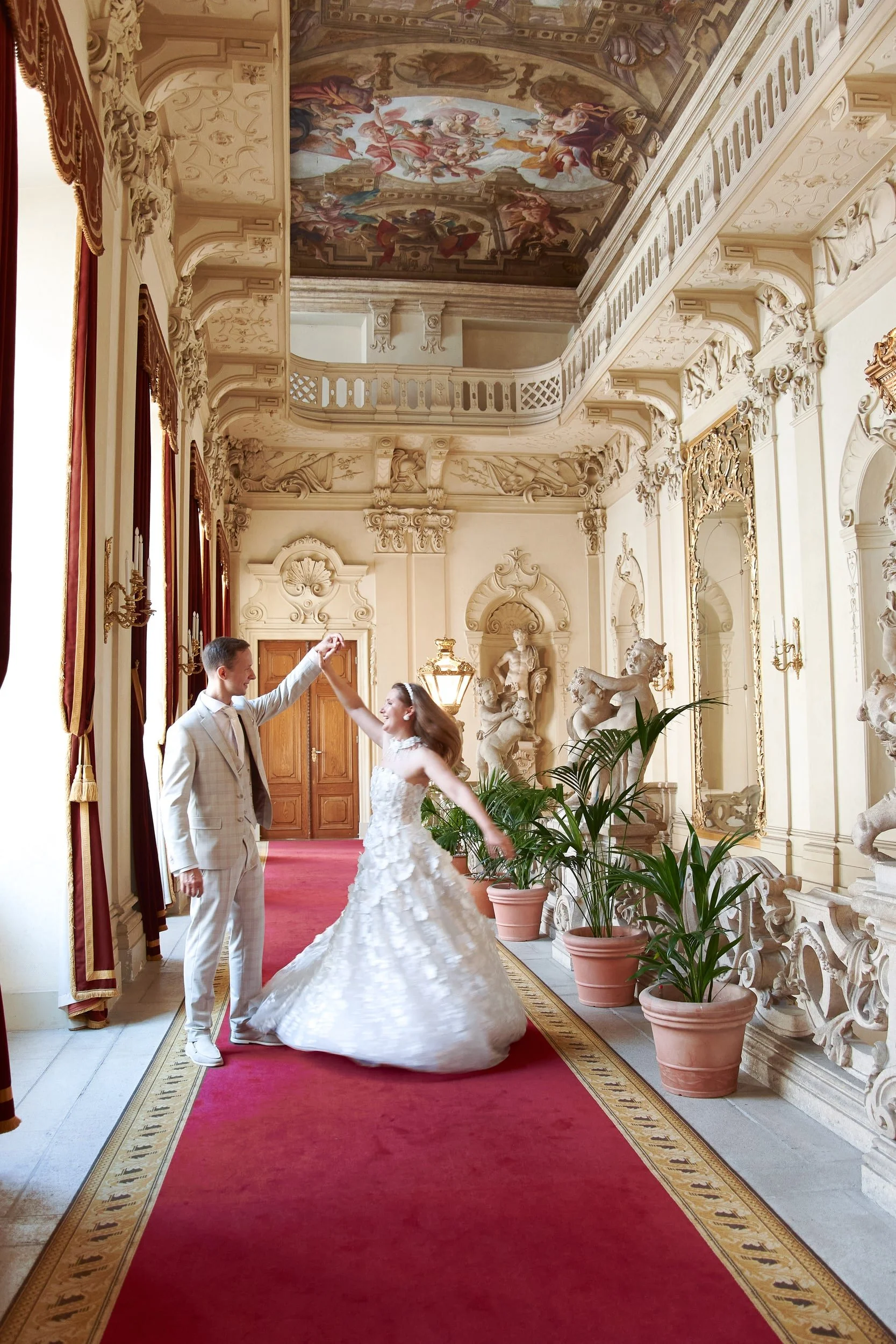 A couple dancing in a luxurious, ornate room with classical sculptures, lush curtains, and decorative molding. The woman wears a white wedding gown, and the man is in a light-colored suit. They are holding hands, and the woman is spinning.