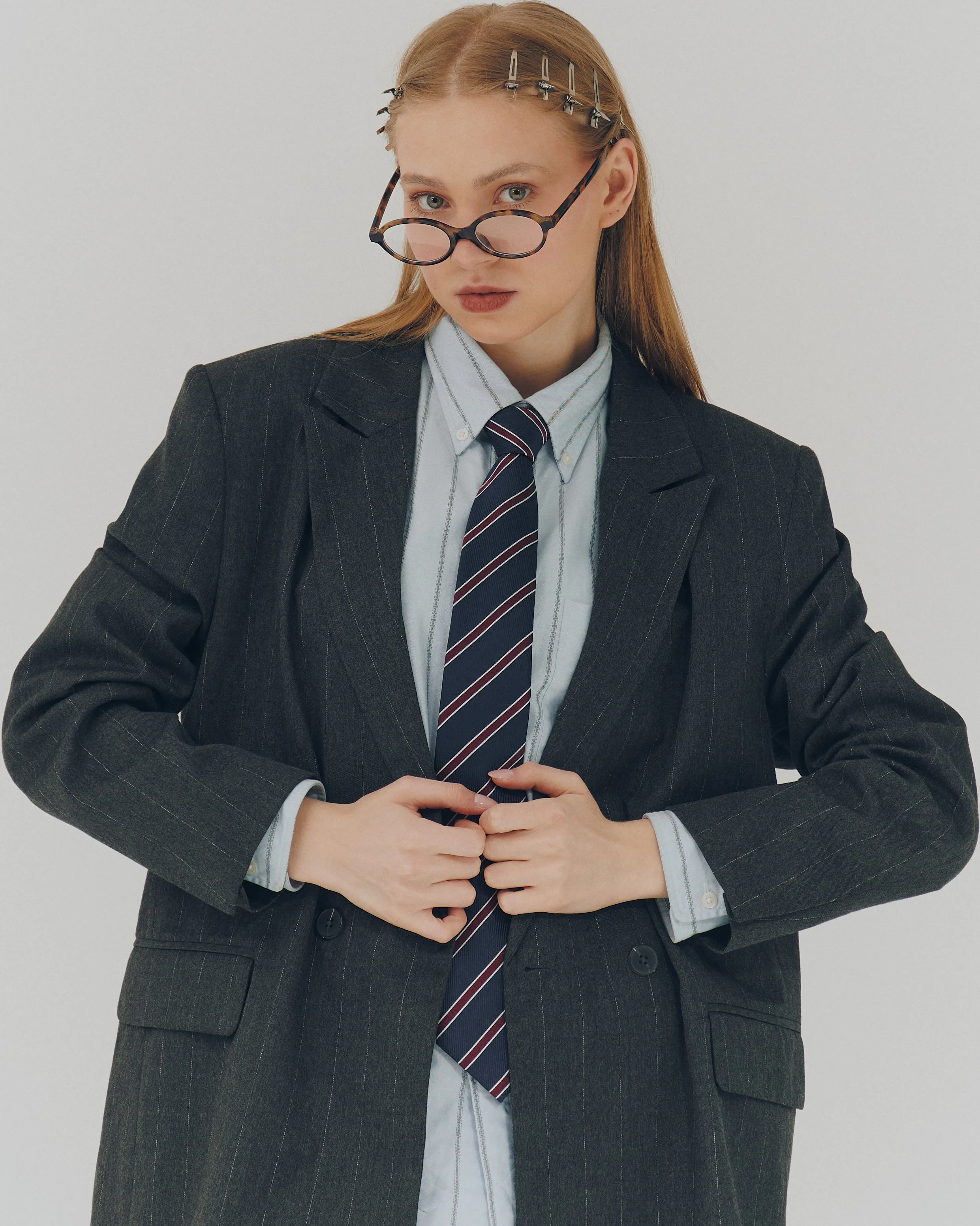 A woman with red hair, wearing glasses on her nose, a dark pinstripe suit, a white collared shirt, and a striped tie, standing against a plain light background.