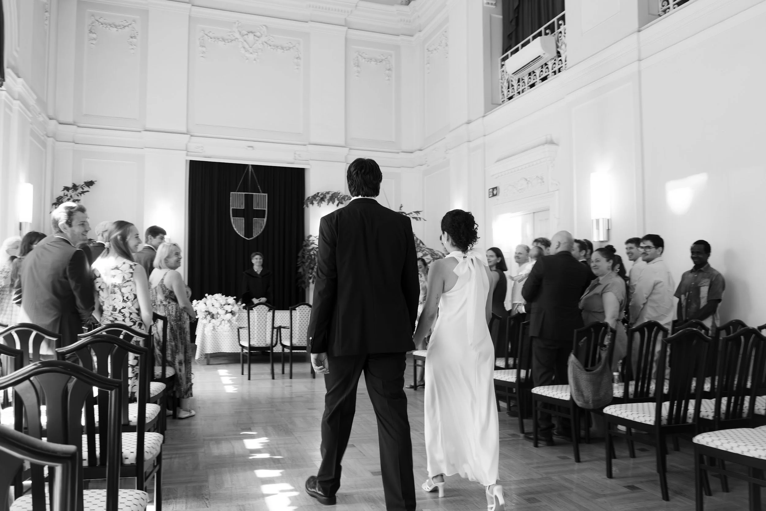 A wedding ceremony taking place in an elegant, high-ceilinged room decorated with classical moldings. The bride and groom are walking down the aisle surrounded by seated guests who are smiling and watching.