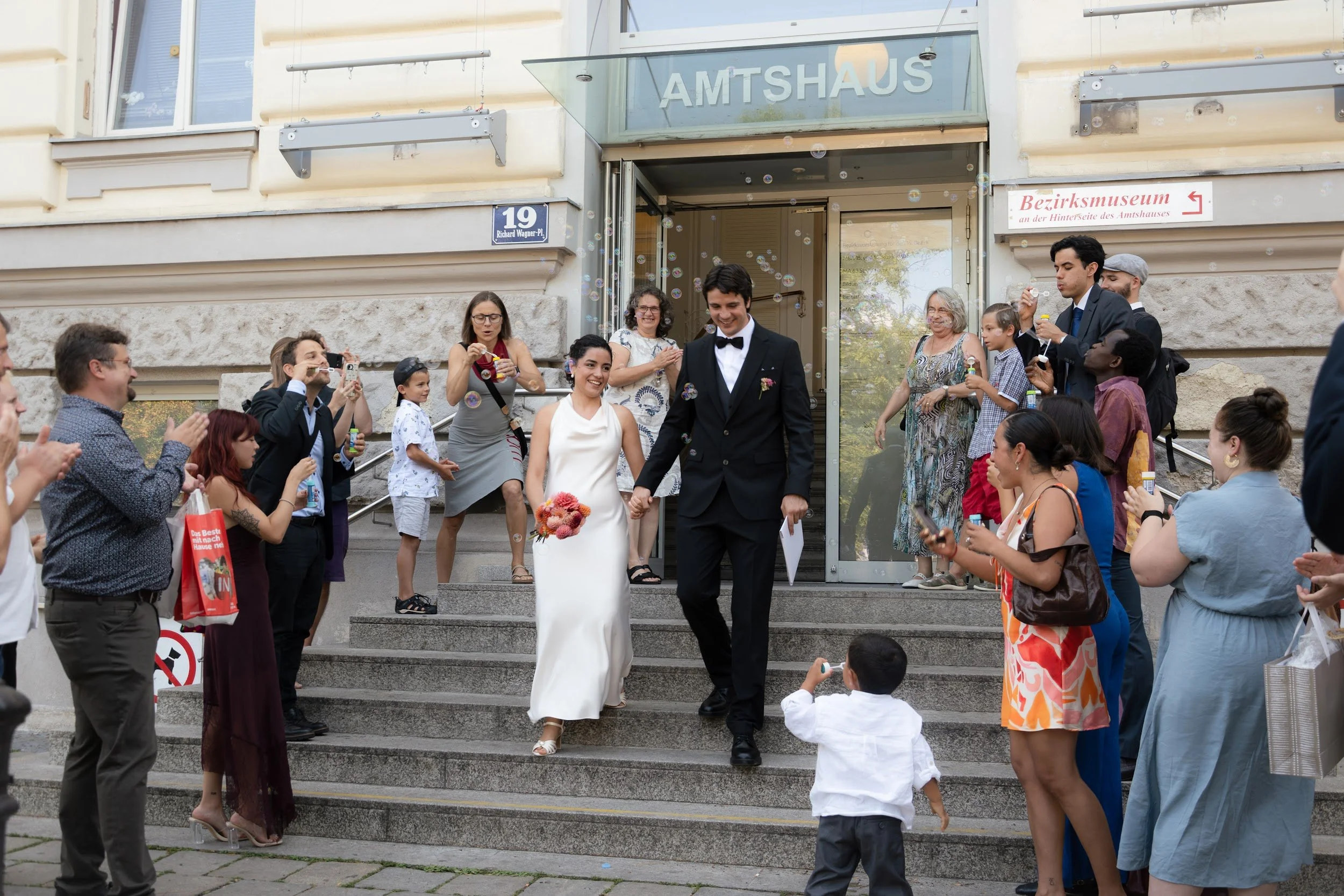 A wedding celebration scene with a bride and groom walking down the steps of a building as guests gather around taking photos and videos, with some blowing bubbles.