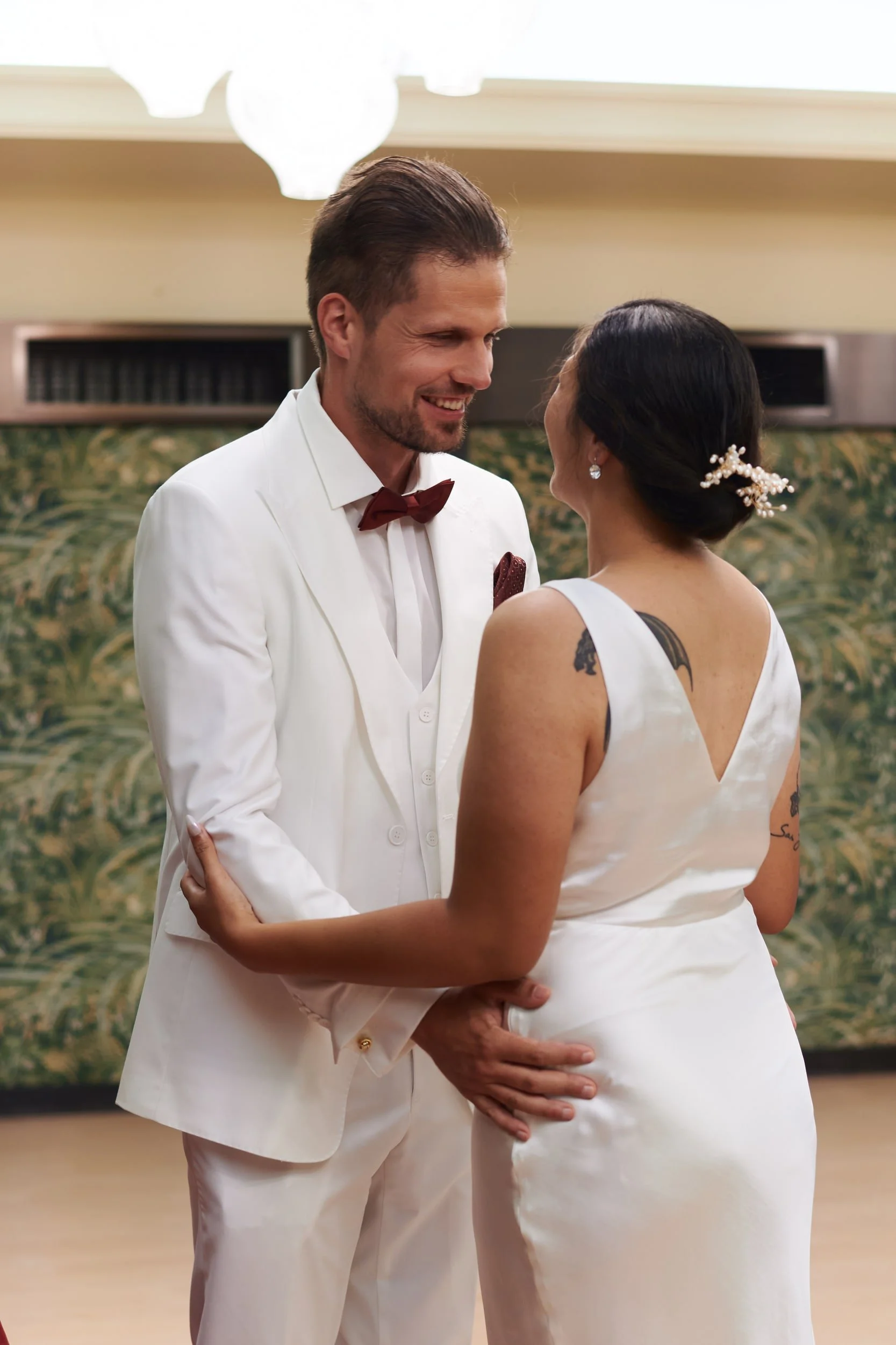 A groom in a white tuxedo with a burgundy bow tie is holding hands and looking at a bride in a white satin wedding dress with a tattoo on her arm, both smiling and sharing an intimate moment in a decorated indoor setting.