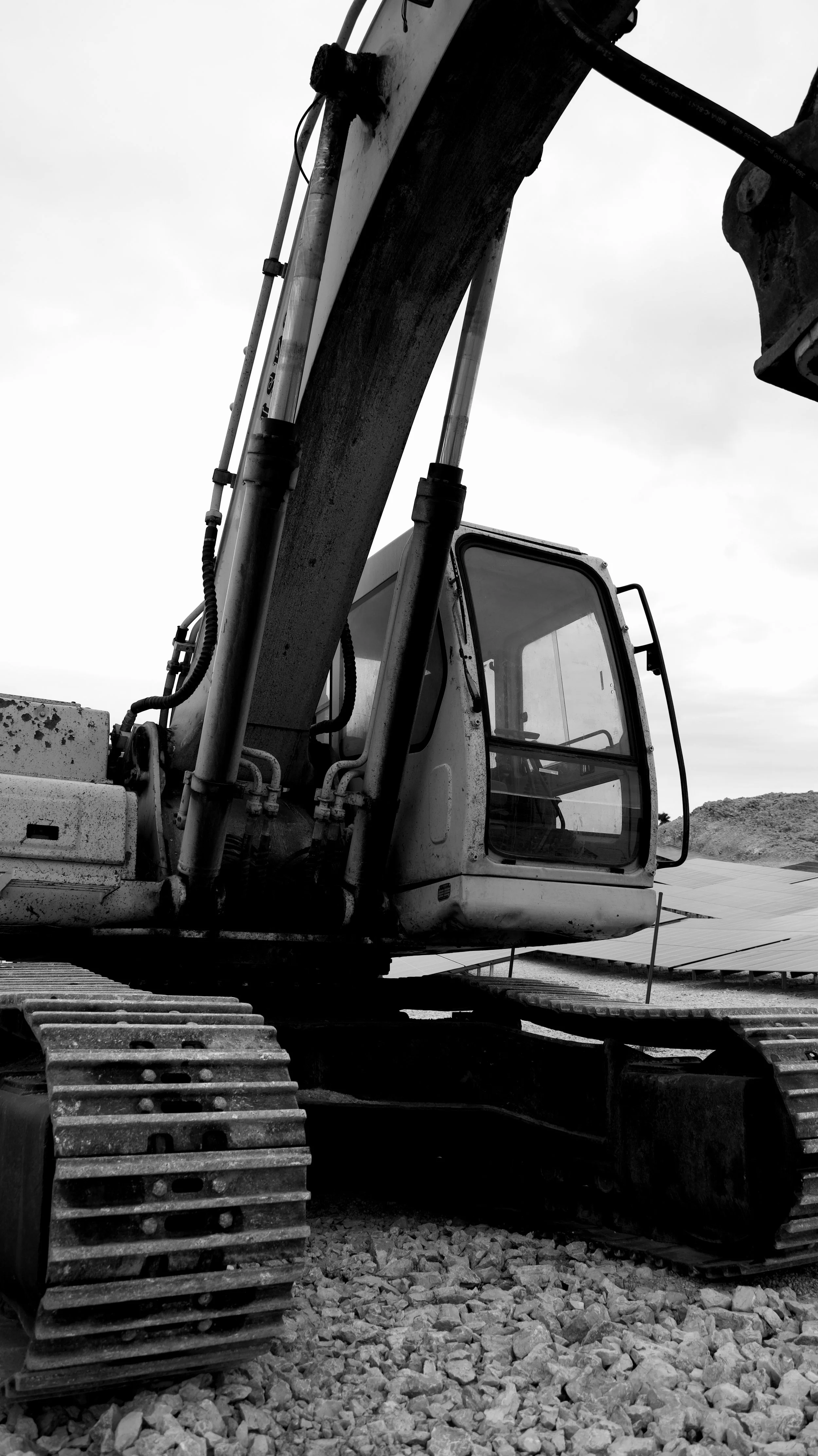 A black and white photo of an excavator on a construction site, with tracks on rocky ground and an overcast sky in the background.