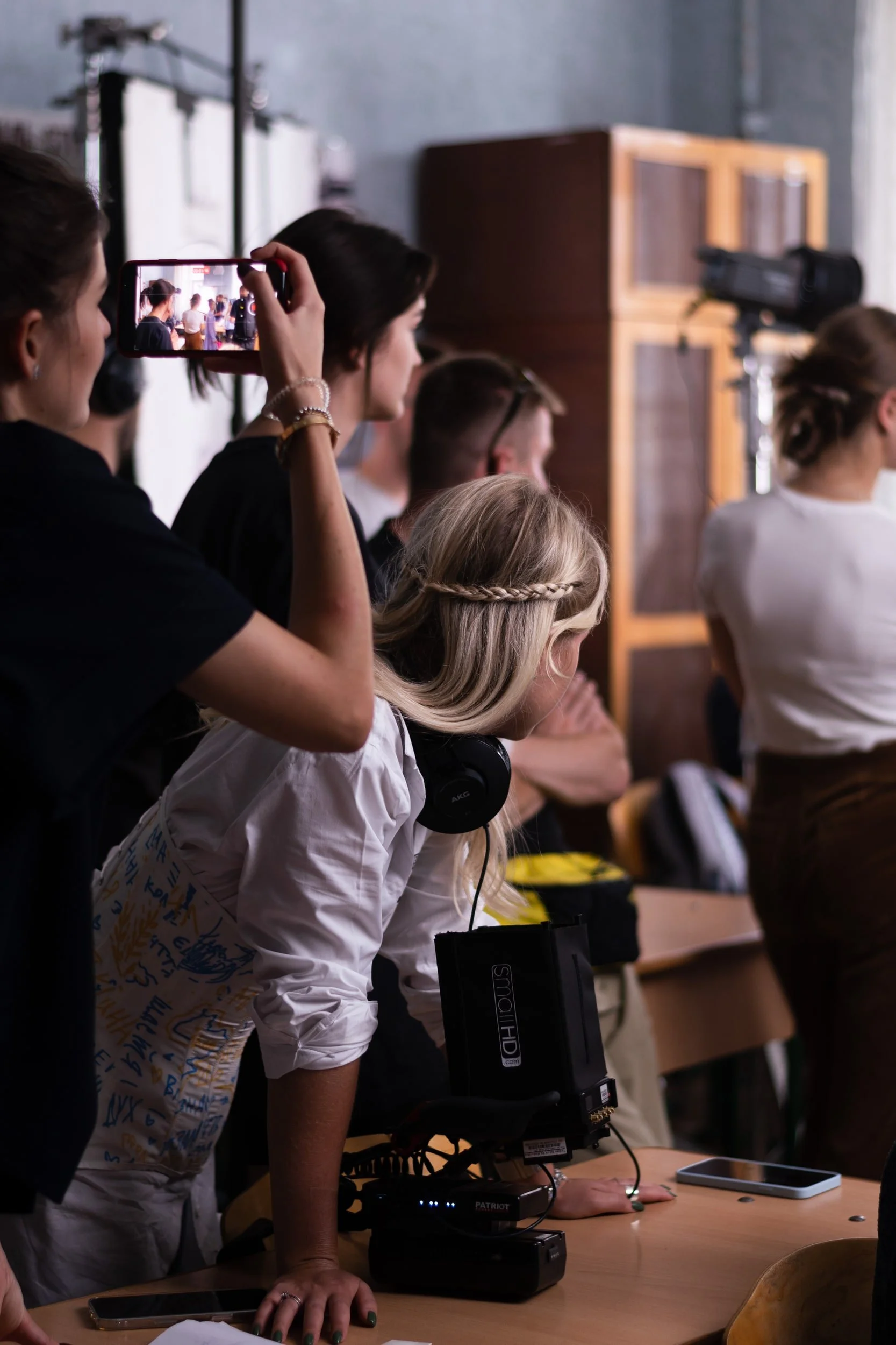 People attending a conference or presentation, with one person taking a photo or video of the event on their phone. The setting appears to be indoors with a wooden cabinet and a camera on a tripod visible in the background.