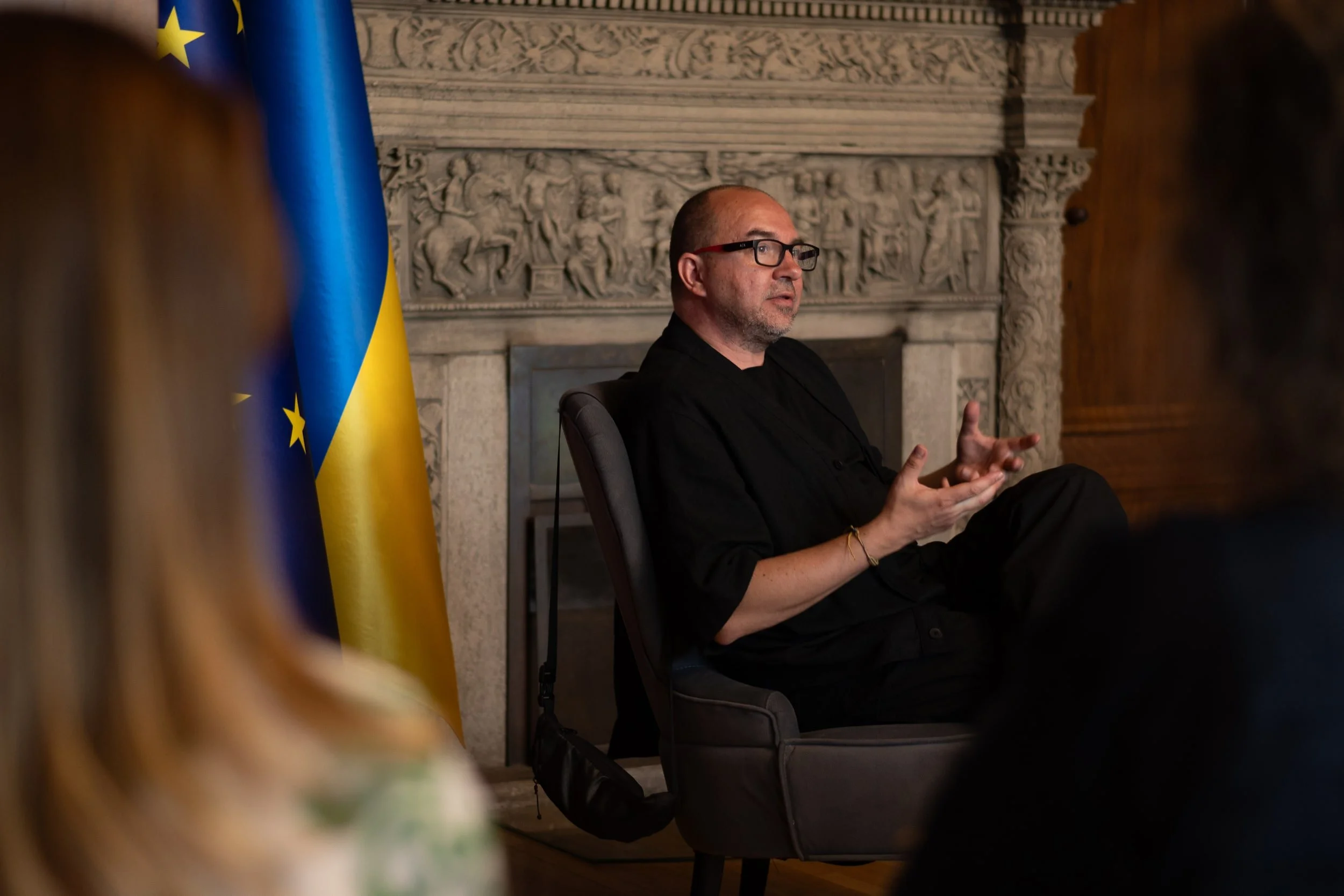 A man wearing glasses, dressed in black, seated in front of a decorative stone wall, speaking or gesturing with hands. Behind him, there are flags, including the European Union flag, visible on the left.