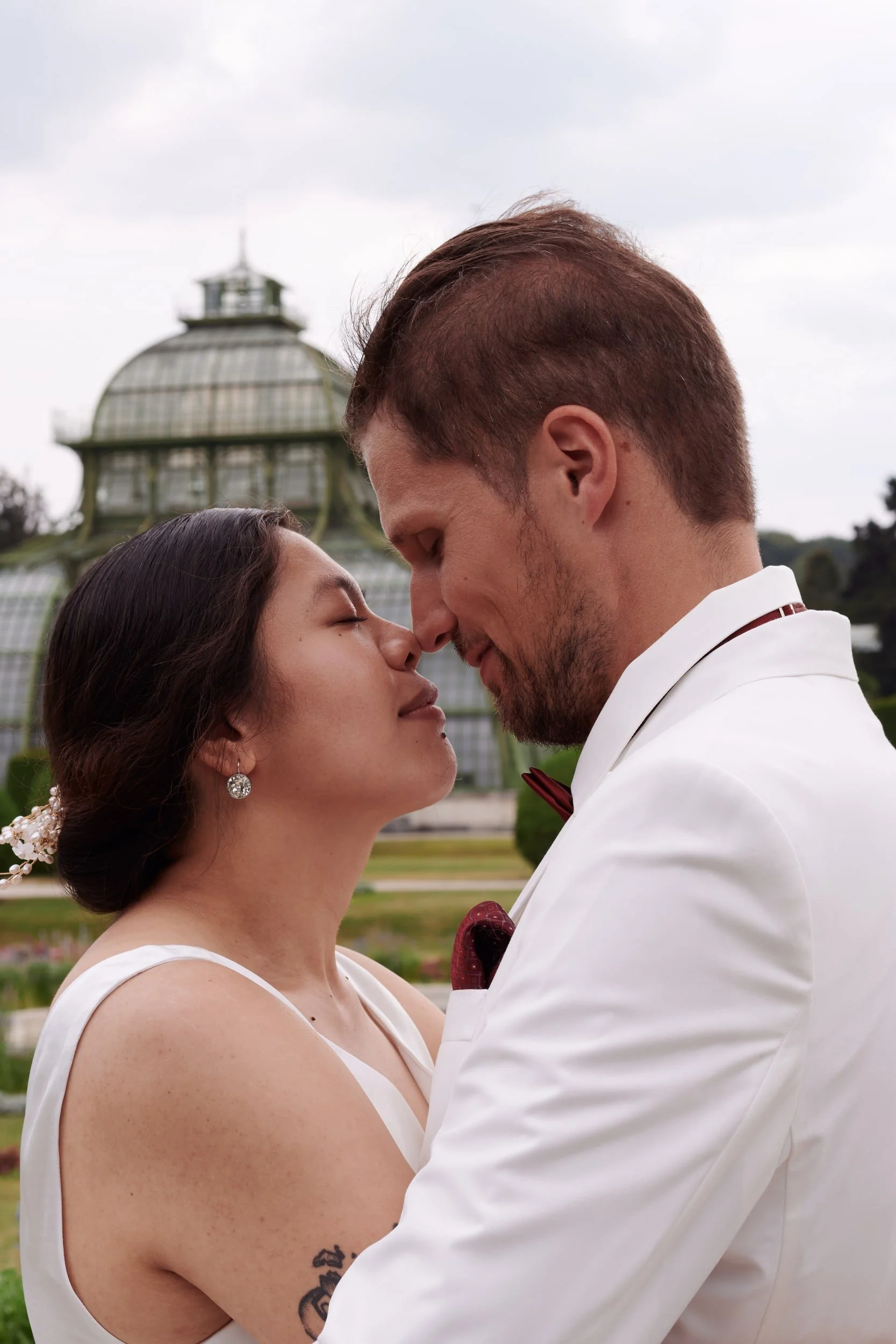 A couple, a man and a woman, are about to kiss outdoors, with their noses touching and eyes closed. The woman wears earrings and a white dress, while the man wears a white tuxedo and a red bow tie. In the background, there is a dome-shaped glass cons