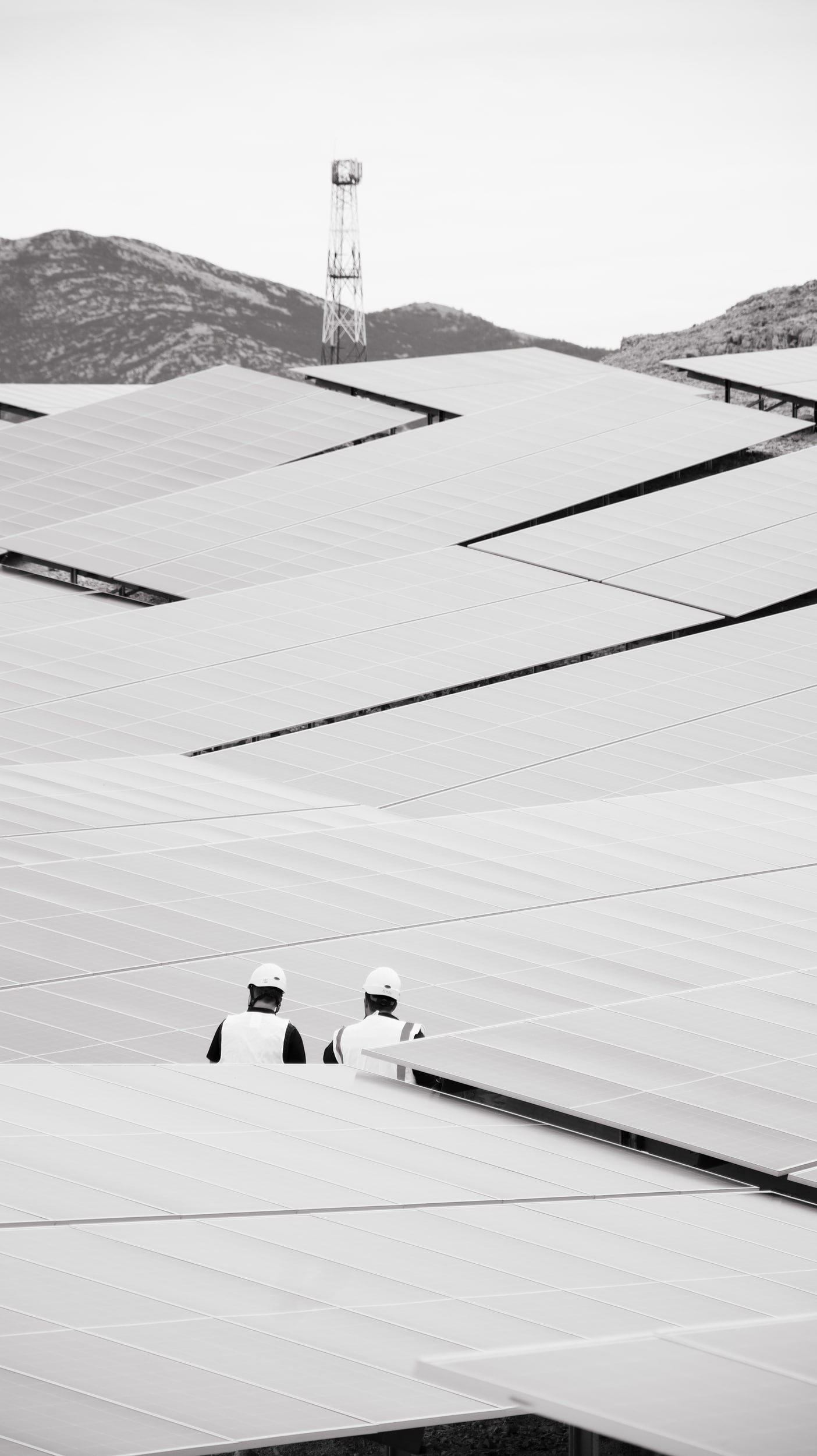 Two workers in safety helmets standing on a large solar panel installation against a mountainous landscape and a tower in the background.