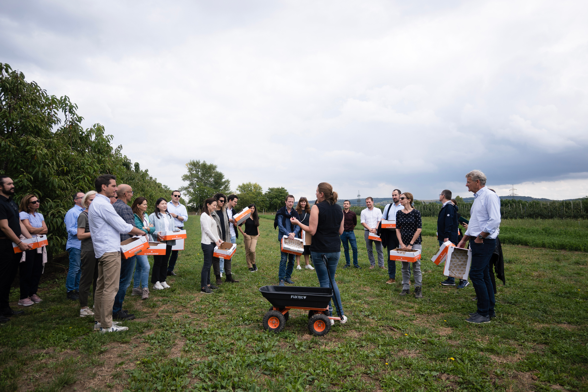 A group of people standing outdoors on a cloudy day, participating in an event or gathering, with some holding boxes and a woman speaking to them.