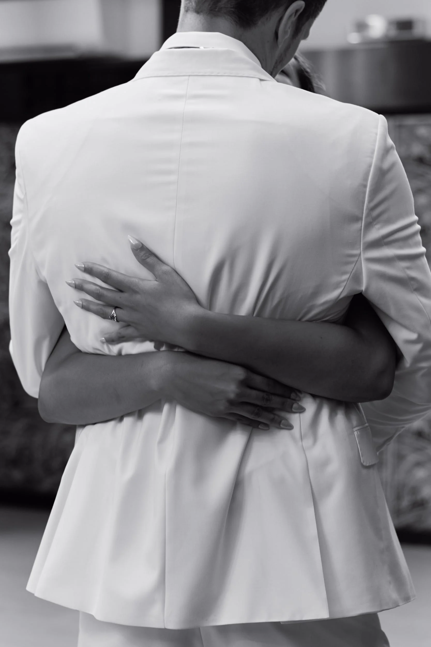 Black and white photo of a person in a light-colored blazer with their back to the camera, hand on another person's waist, and possibly dancing or embracing.