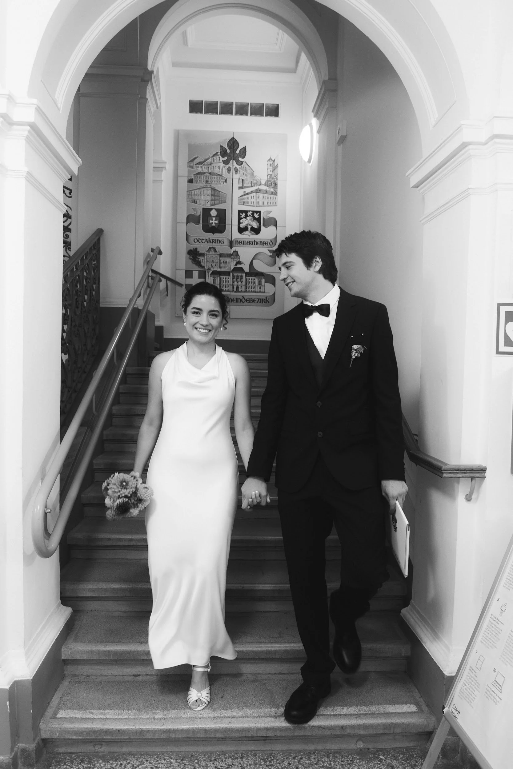 Black and white photo of a smiling woman in a white dress and a man in a tuxedo holding hands, standing on staircase inside a building with a large painting or banner behind them.