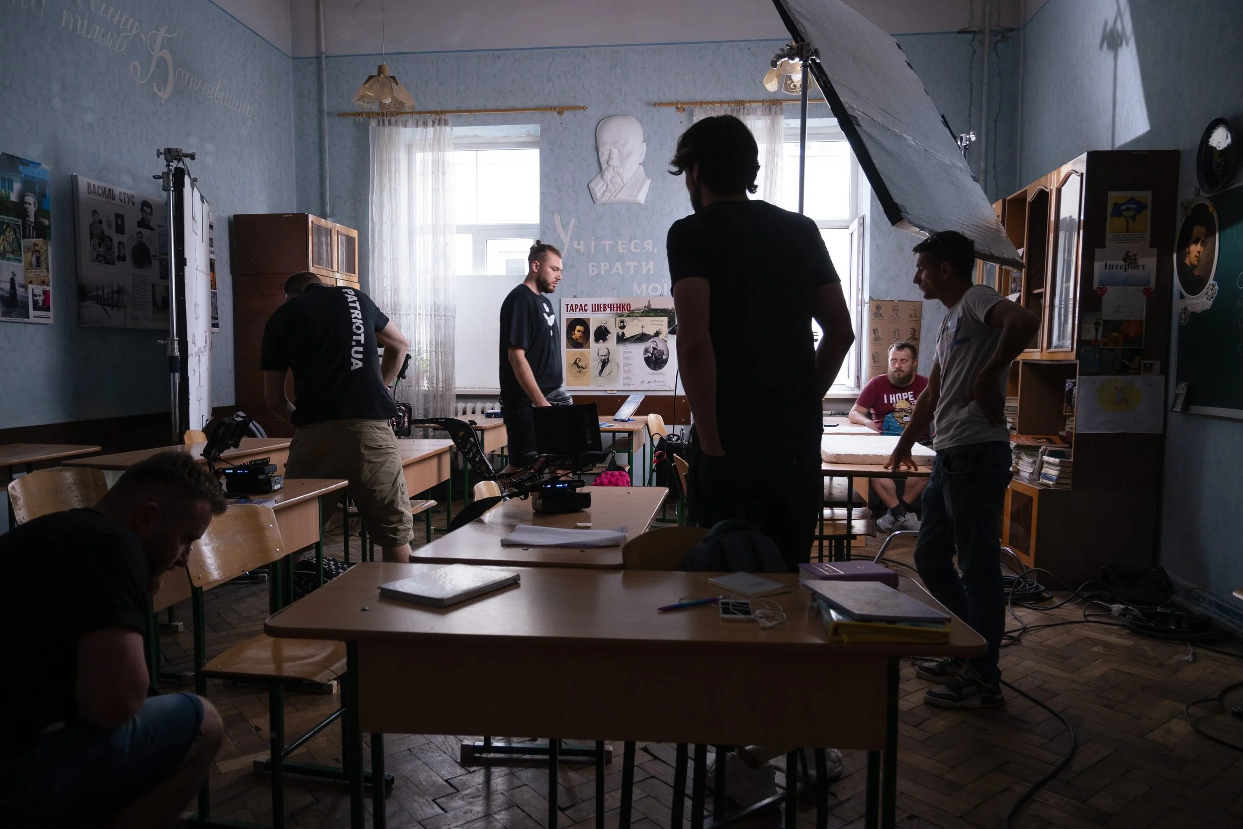 A group of people in a classroom setting with photography and lighting equipment, some standing and others sitting at desks, engaged in a discussion or preparing for a scene.