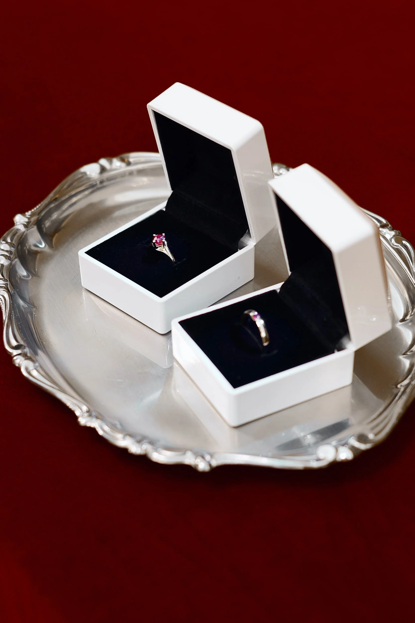 A silver tray holds two open jewelry boxes with rings inside, one with a pink gemstone, on a dark red surface.