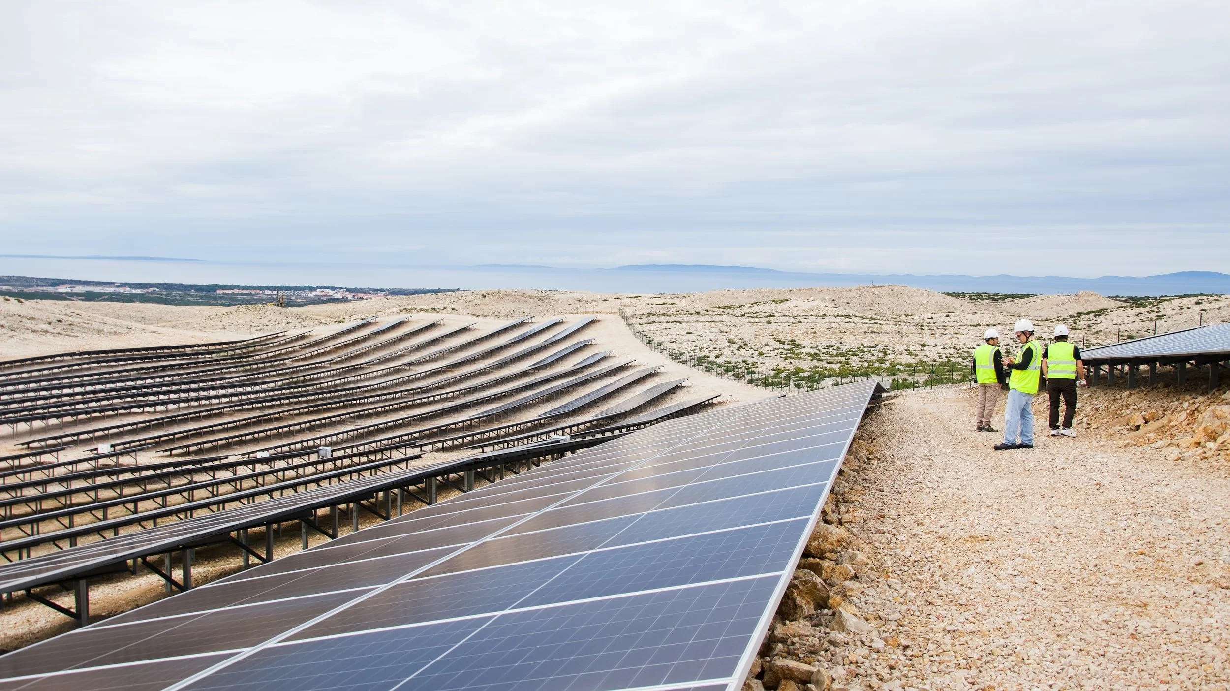 A solar farm under construction with workers wearing safety vests and helmets inspecting the installation of solar panels on a hillside with a cloudy sky in the background.