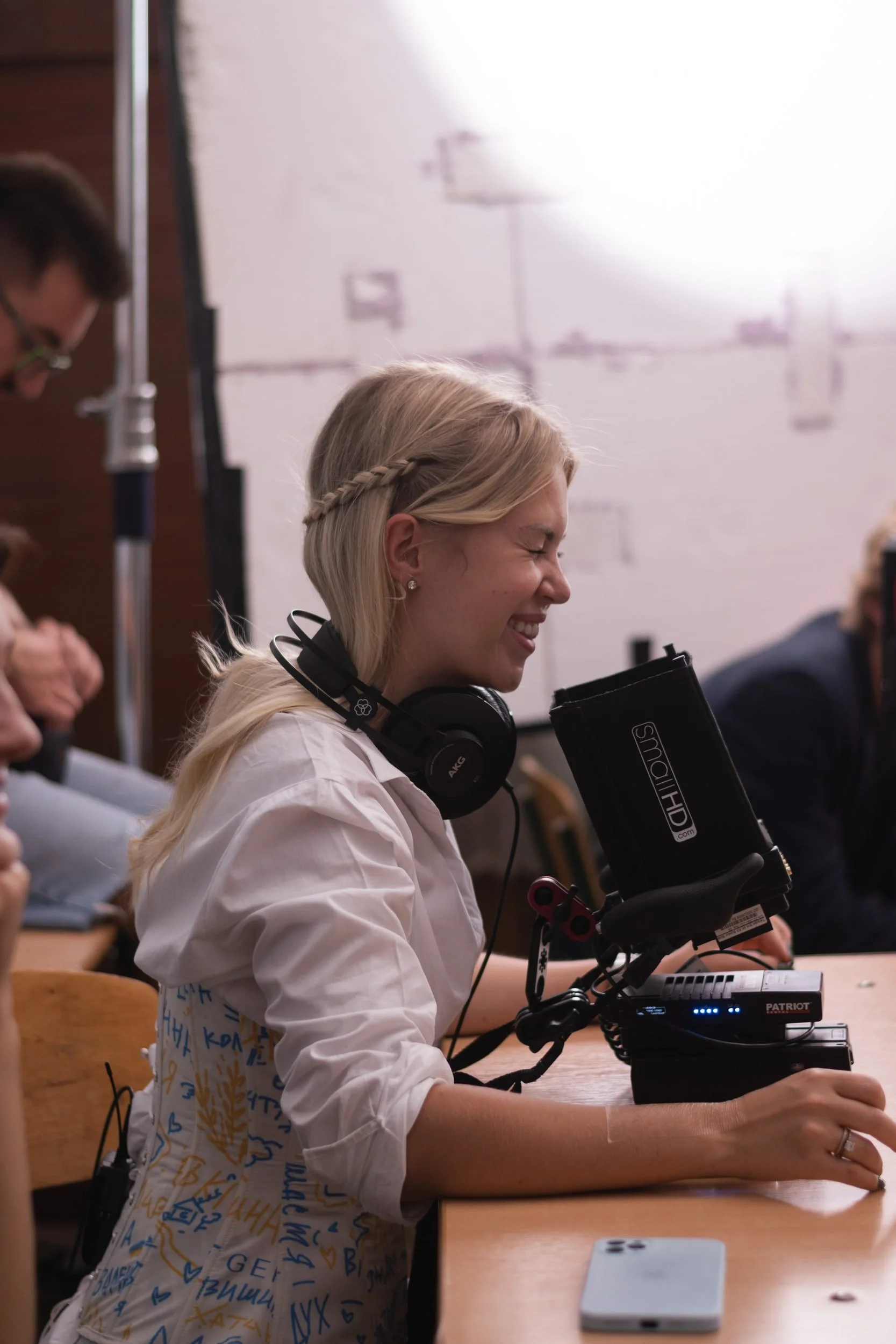 A woman with blonde hair in a braid smiling while sitting at a desk using a microscope in a classroom or lab setting.