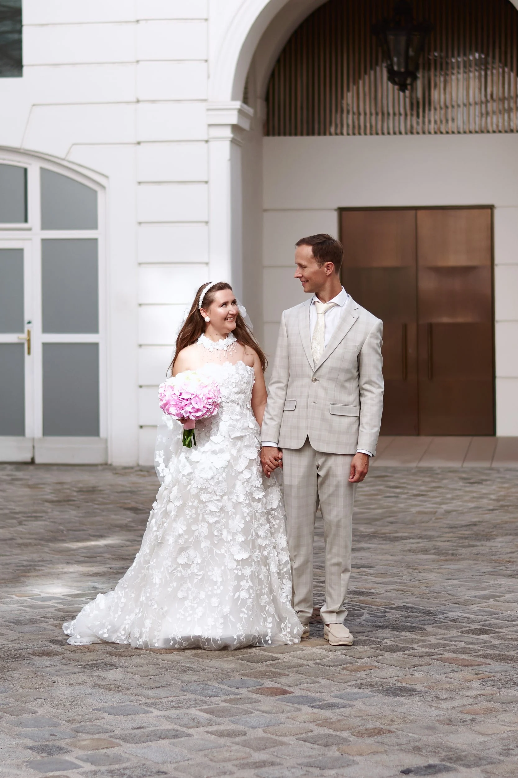 A bride and groom are holding hands outside a white building with large windows and an arched entrance. The bride is wearing a white lace wedding dress and holding a pink bouquet, while the groom is in a light-colored plaid suit. They are smiling at 